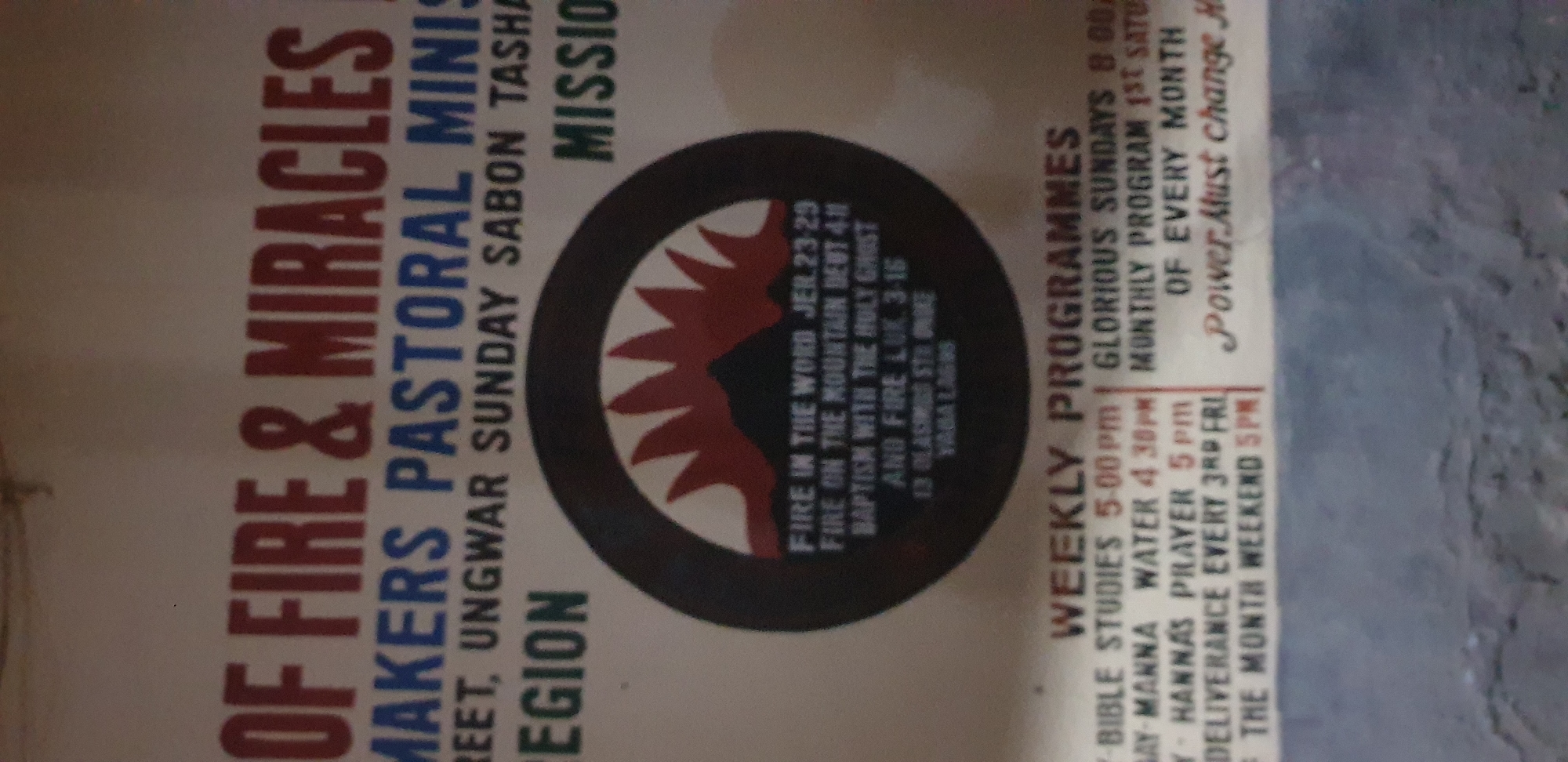 The image is a photograph of a poster that appears to be displayed outdoors, possibly on a wall. The poster advertises a religious organization called "FIRE & MIRACLES MAKERS PASTORAL MINIS" located in "REGION" which is identified as Narayi, Nigeria.
The poster has several sections of text. The top portion prominently displays "OF FIRE & MIRACLES" in large red letters, followed by "MAKERS PASTORAL MINIS" in blue. Below this, there are lines of text that are partially obscured or difficult to read, mentioning "STREET, UNGWAR SUNDAY SABON TASHA" and "REGION". To the right, "MISSIO" is visible, suggesting "MISSION".
A circular graphic in the center of the poster depicts a mountain with flames erupting from its peak, rendered in red against a dark background. Inside the circle, there is text that reads: "FIRE IN THE WORD JER. 23-29", "FIRE ON THE MOUNTAIN DEUT. 4:11", "BAPTISM WITH THE HOLY GHOST AND FIRE LUK. 3-16", "13 OLASIMIDI STH DUKE YABA LAGOS". This suggests that although the primary location is Narayi, Nigeria, there may be a secondary address or reference in Yaba, Lagos.
The lower part of the poster lists "WEEKLY PROGRAMMES". This section includes:
- "BIBLE STUDIES 5:00pm"
- "-MANNA WATER 4.30pm"
- "- HANNAS PRAYER 5pm"
- "- DELIVERANCE EVERY 3RD FRI"
- "- OF THE MONTH WEEKEND 5PM"
To the right of the weekly programs, "GLORIOUS SUNDAYS 8 00a" and "MONTHLY PROGRAM 1ST SATU" are listed, followed by "OF EVERY MONTH". The phrase "Power Must change H" is also visible in a more stylized font.
The overall poster appears to be printed on a light-colored background, likely paper. The lighting suggests it is daytime. The lower portion of the image shows a textured surface, possibly a concrete wall or pavement, below the poster. There are no people visible in the image.