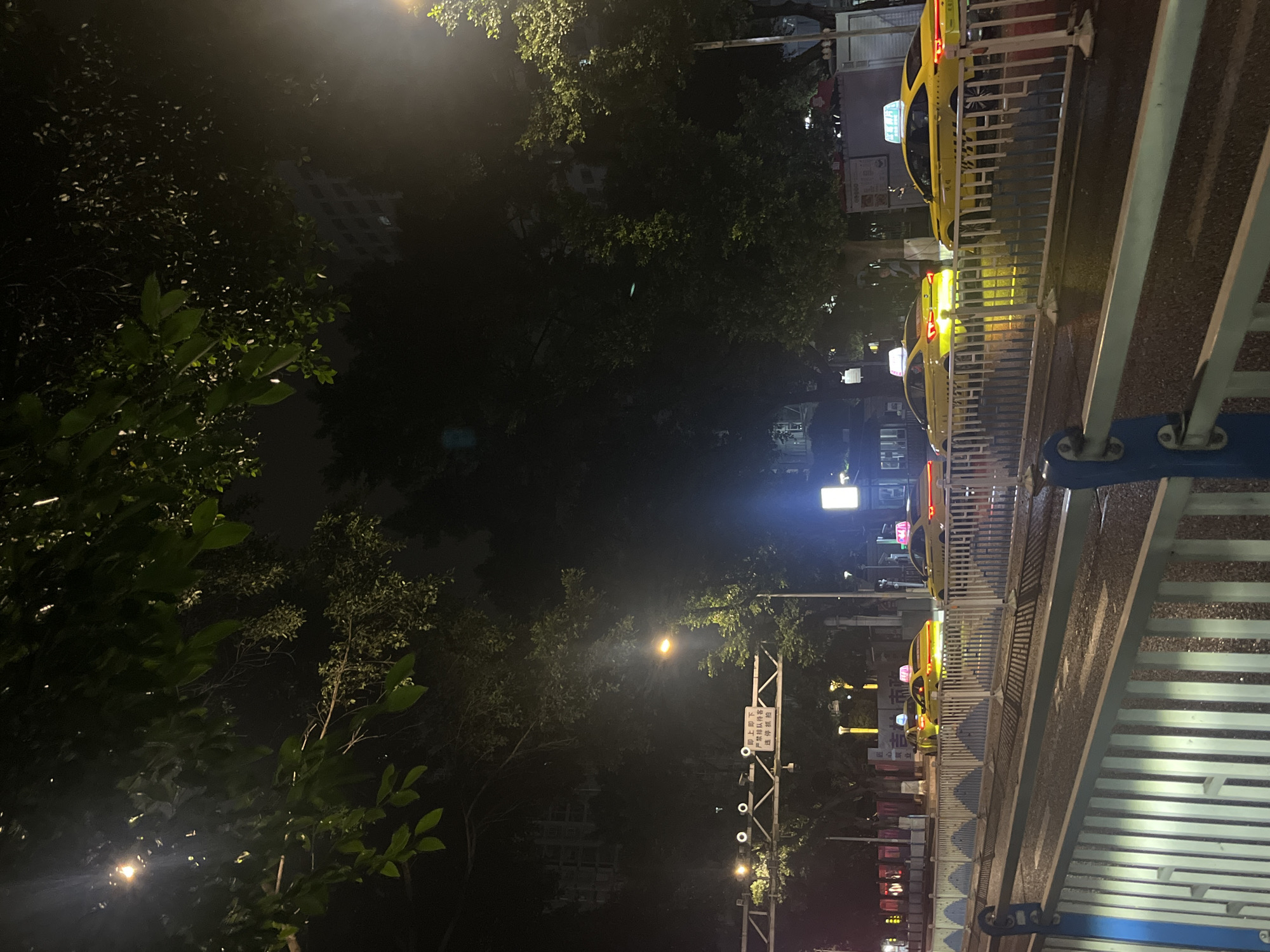 The image depicts an urban street scene at night in Jiulongpo District, China. In the immediate foreground, a multi-tiered metal railing system is present. The lower section features a blue top rail with white vertical slats, while the upper section has white vertical slats with blue geometric patterns. Beyond this railing, the asphalt road surface is wet, reflecting various light sources.
A line of yellow sedan-style taxis occupies the road. From right to left: one taxi is partially visible with illuminated red taillights; two subsequent taxis are brightly lit, featuring yellow bodies and illuminated white or blue roof lights; and a fourth taxi is partially visible further down the street.
An overhead gantry structure spans the road, equipped with multiple circular lights and a white rectangular sign. The sign displays Chinese characters: "即上即下 严禁排队候客 违停抓拍," which translates to "Pick up and drop off immediately. Queueing for passengers is strictly prohibited. Illegal parking will be photographed."
Streetlights on poles emit a warm, orange-yellow light, illuminating portions of the road and the surrounding environment. Dense green foliage from numerous trees dominates the upper and side sections of the image, partially obscuring multi-story buildings in the background. The sky is dark. A single, indistinct human figure is faintly visible standing on the sidewalk behind the taxis, between two trees. No specific actions or interactions are discernible.