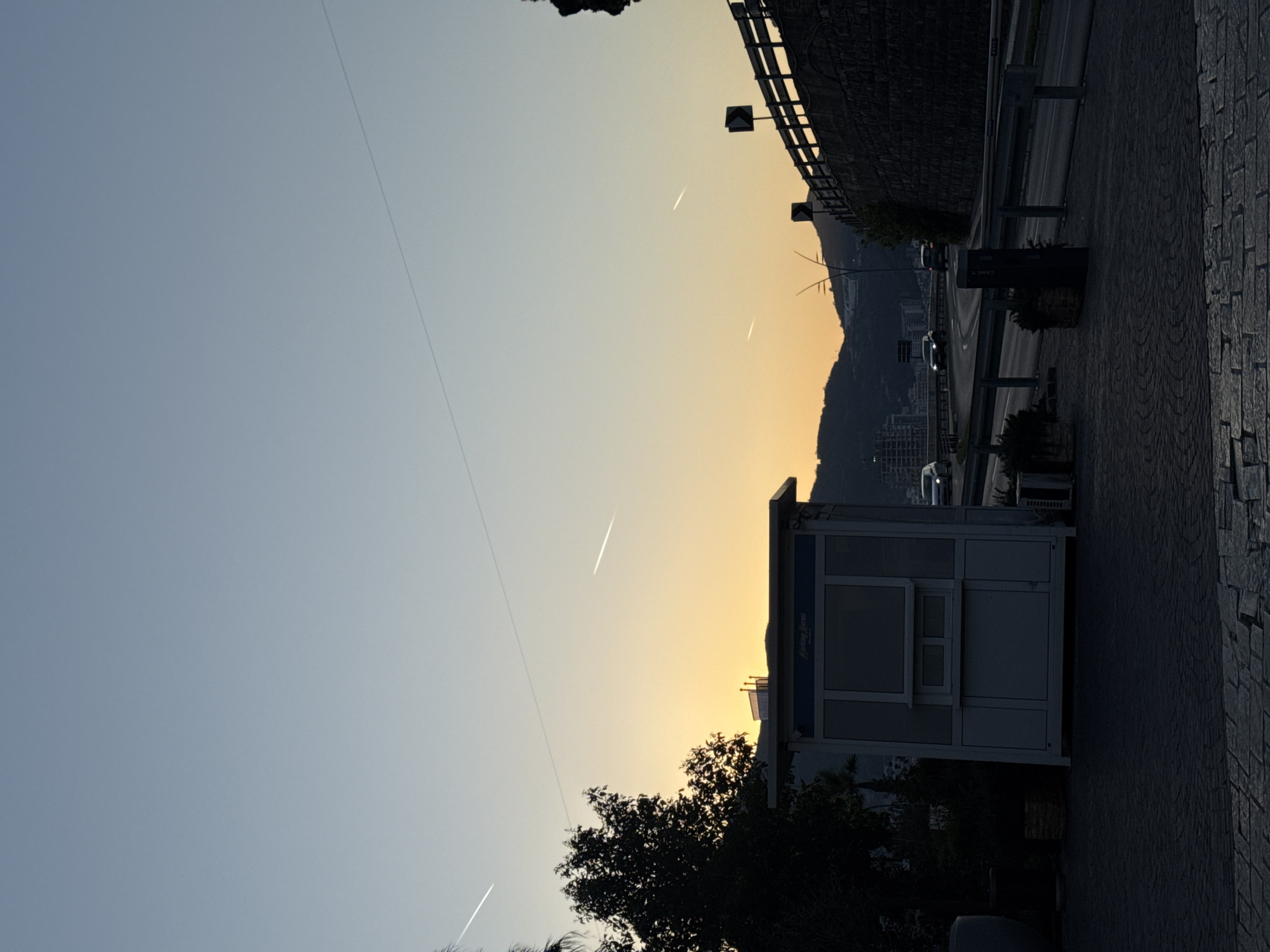 The image shows a twilight scene in an urban environment. In the foreground, a white structure with windows, possibly a kiosk or small building, stands on a paved walkway. Adjacent to the walkway is a road with passing vehicles. The sky transitions from blue to orange towards the horizon. A mountain or hill is visible in the background, partially obscured by the glow of the setting sun. The location is the city of Unknown, Unknown.