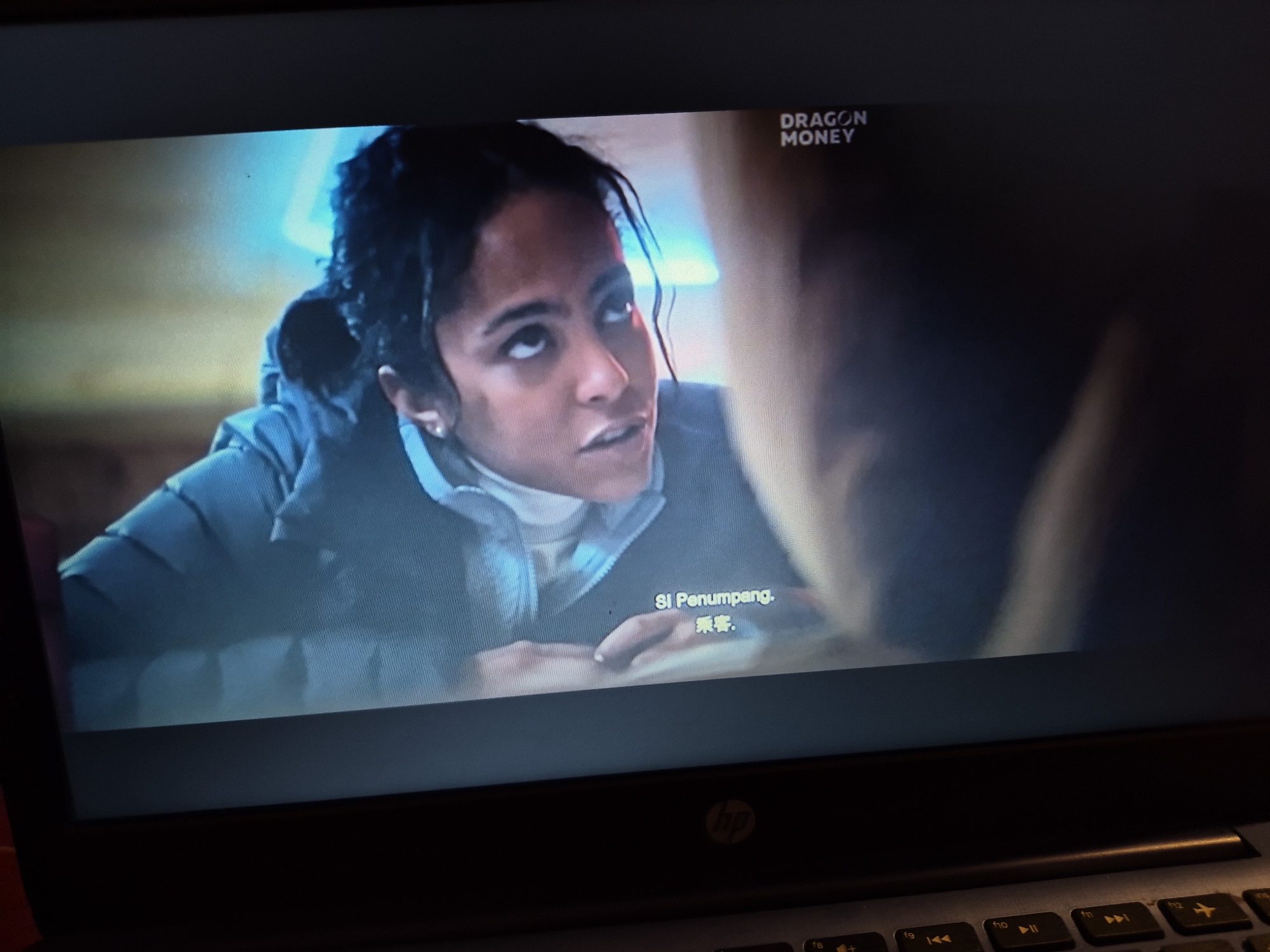 The image shows a close-up of a laptop screen displaying a scene from a movie or television show. In the foreground, a woman with dark hair pulled back is looking upwards and to the left with a surprised or questioning expression. She is wearing a light-colored turtleneck sweater and a dark blue puffer jacket. In the top right corner of the screen, the text "DRAGON MONEY" is visible. Below the woman, there is text in a foreign language, appearing to be "SI Penumpang." and some Chinese characters. The background is blurred, with hints of blue light, suggesting an indoor setting. The laptop itself is visible at the bottom of the frame, with a dark keyboard and a HP logo on the right side. There is no indication of the image being located in Sandwell, United Kingdom.