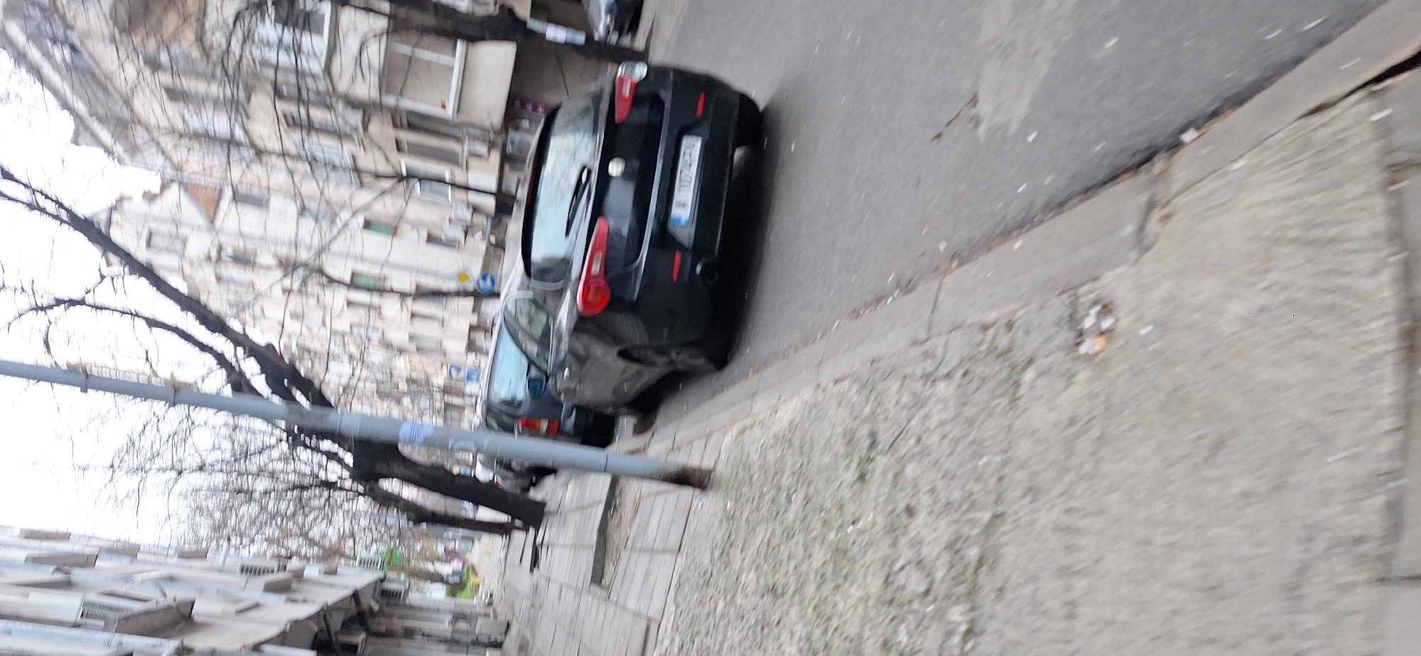 The image shows a street scene in Burgas, Bulgaria. A black car is parked along the side of the road, and a smaller car is parked behind it. Bare tree branches are visible on the left of the frame. A streetlamp stands near the sidewalk and a building is visible in the background. The sidewalk is made of concrete and slightly dirty. The license plate of the black car is partly visible.