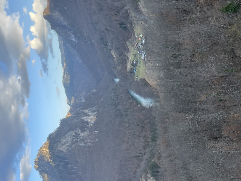 The image presents an expansive aerial view of the Tara River Canyon near Đurđevića Tara, Montenegro. The scene is dominated by rugged, steep mountainsides flanking a deep valley. These slopes are extensively covered in dense, bare deciduous forest, indicating a late autumn or winter season, with some rocky outcrops visible on higher elevations. The peak of the mountain on the far left is partially illuminated by direct sunlight, while other areas are in shadow or diffuse light.
A clear, light blue river, presumed to be the Tara River, winds through the canyon floor, flowing from the mid-left towards the central-right of the frame. On the right side of the river, nestled on a patch of green grassland within the valley, a small, isolated settlement is visible. This cluster comprises approximately five distinct buildings with varying roof colors (reddish-brown, dark grey, light blue) and several smaller auxiliary structures. The immediate foreground consists of a dense canopy of bare tree branches. The upper third of the image reveals a partially cloudy sky, with patches of clear blue interspersed with scattered white and grey clouds. No individuals, vehicles, or specific activities are discernible.