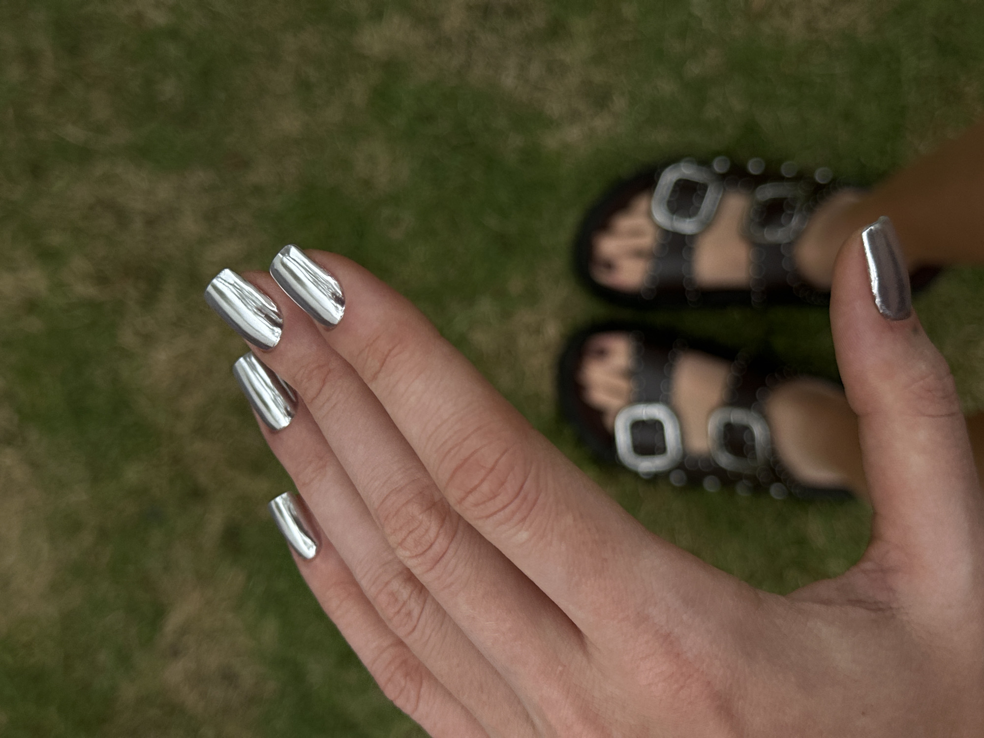 The image shows a close-up of a hand with silver chrome nail polish, positioned in front of a pair of black leather sandals with silver buckles. The background consists of blurred green grass, which suggests the location may be outdoors. While it's impossible to precisely confirm the location in Dalung, Indonesia, without additional context, the presence of grass and sandals could indicate a casual setting. The focus of the image is clearly on the hand and the shiny nails, highlighting a beauty or fashion aspect.