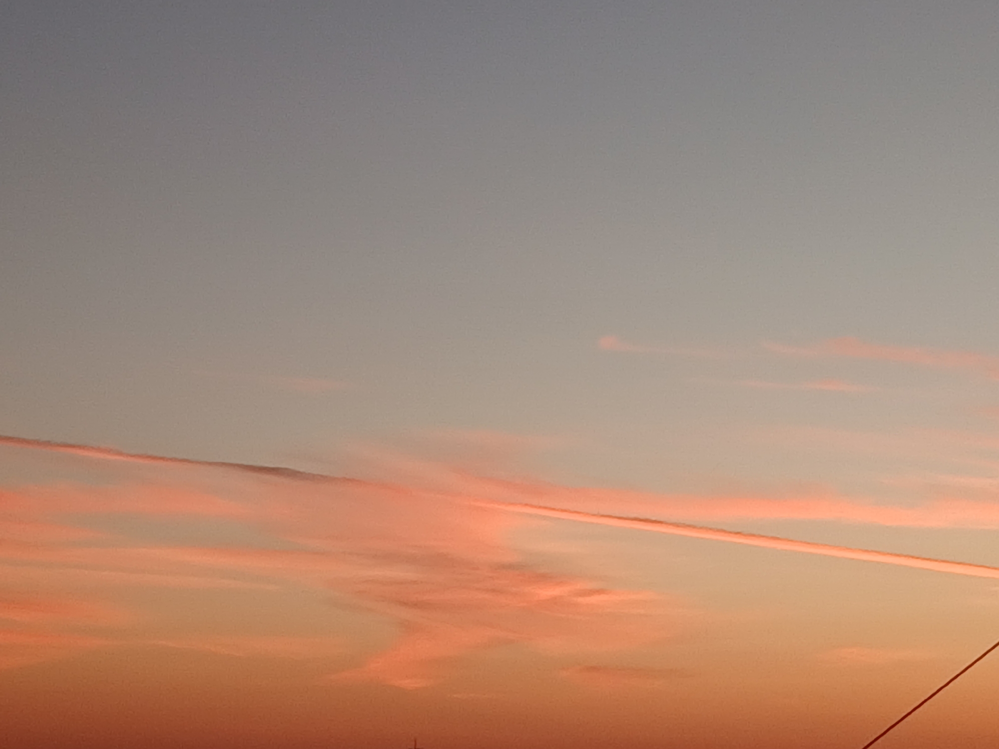 The image depicts a sky with a gradient of colors, transitioning from a pale blue at the top to a warm orange at the bottom, indicative of a sunset or sunrise. Wisps of pink and orange clouds are scattered across the sky. A prominent contrail, also tinged with pink, stretches diagonally across the frame. The lower portion of the image shows a sliver of the horizon with faint silhouettes of structures, possibly buildings or antennas. The scene appears to be an outdoor environment, likely an open area or cityscape. The image was taken in Makinsk, Kazakhstan.