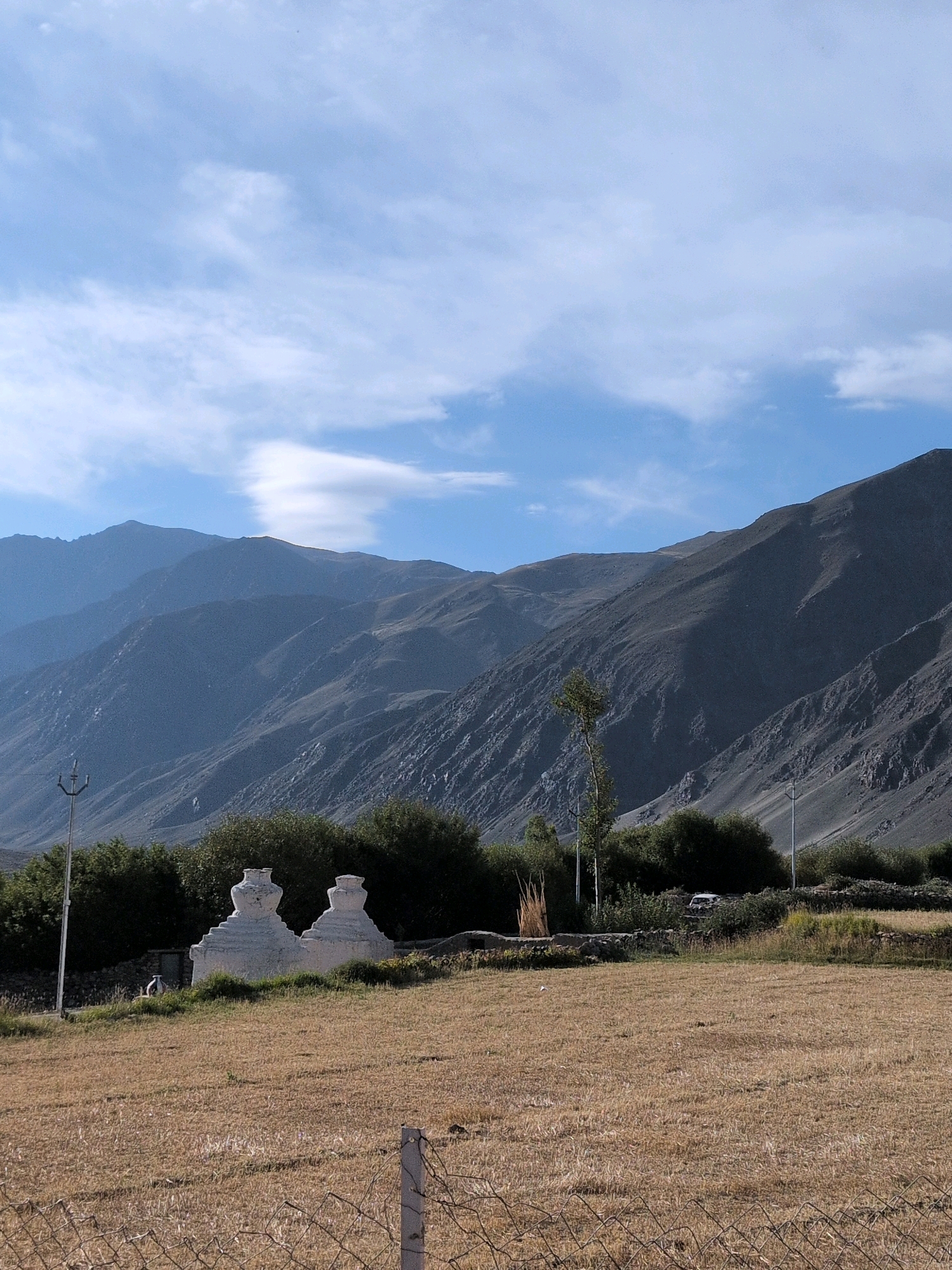 In Skidmang, India, two white stupas stand in a dry, grassy field. Behind the stupas, a line of dark green trees forms a backdrop. Further back, a barren, mountainous landscape dominates the scene, with layered, shadowed peaks stretching towards a pale blue sky dotted with white clouds. A wire fence with a metal post is visible in the foreground. A utility pole with an insulator is situated to the left of the stupas. A person is seated on a low wall behind the stupas.