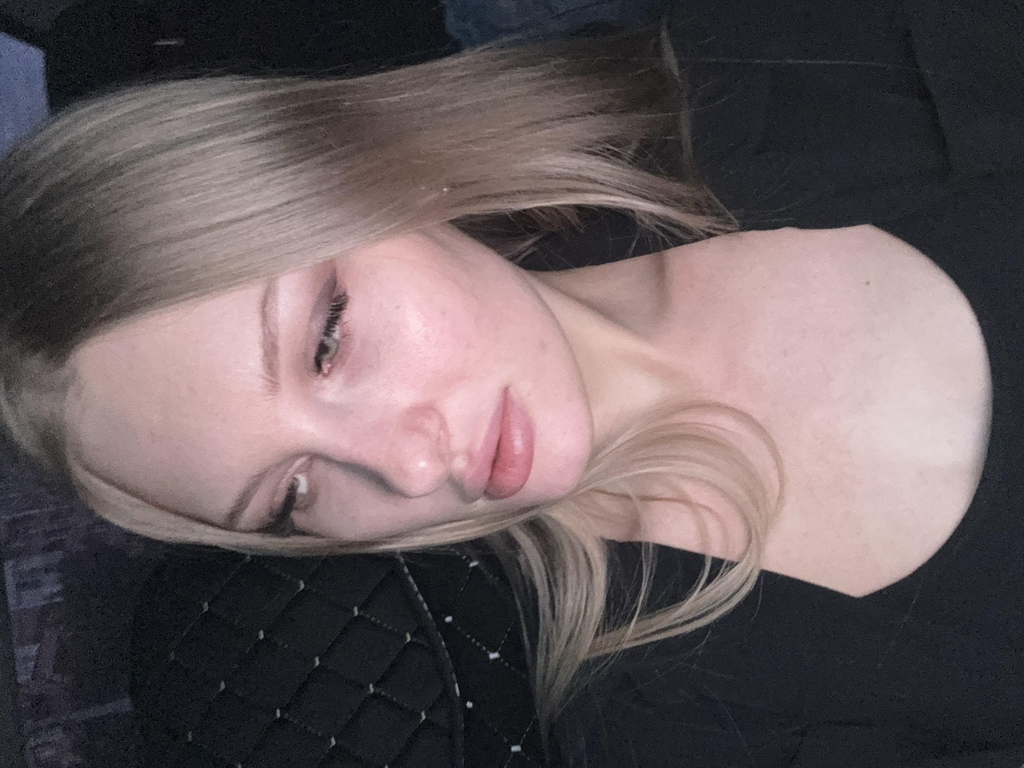 A young woman with blonde hair is lying on a black quilted surface, possibly a cushion or headrest. She is wearing a black top. Her gaze is directed downwards. The location is unknown.