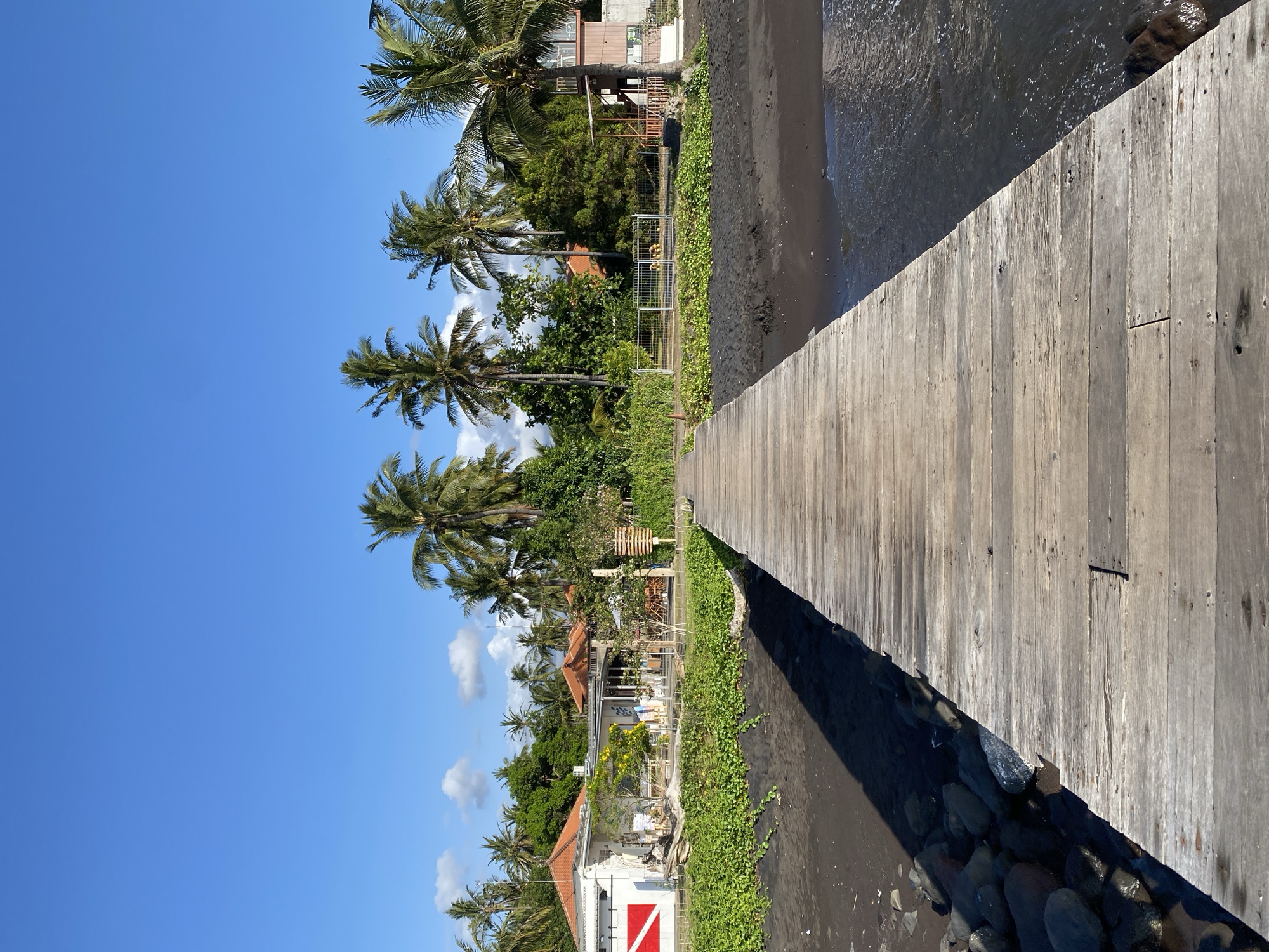 The image depicts a wooden pier extending towards a tropical beach. The pier is made of weathered wooden planks laid horizontally. To the left of the pier, there is black sand with a line of green vegetation. Beyond the vegetation, a row of buildings with orange roofs is visible, interspersed with numerous palm trees. The buildings appear to be part of a resort or residential area. A red and white flag, possibly a dive flag, is visible on the side of one building. To the right of the pier, the ocean gently laps at the shore, with ripples and small waves visible. The sky is a clear, bright blue with a few scattered white clouds. The overall scene suggests a sunny day in a tropical location, likely a beach resort. The location is identified as Bulusan, Indonesia. There are no people or animals visible in the image.