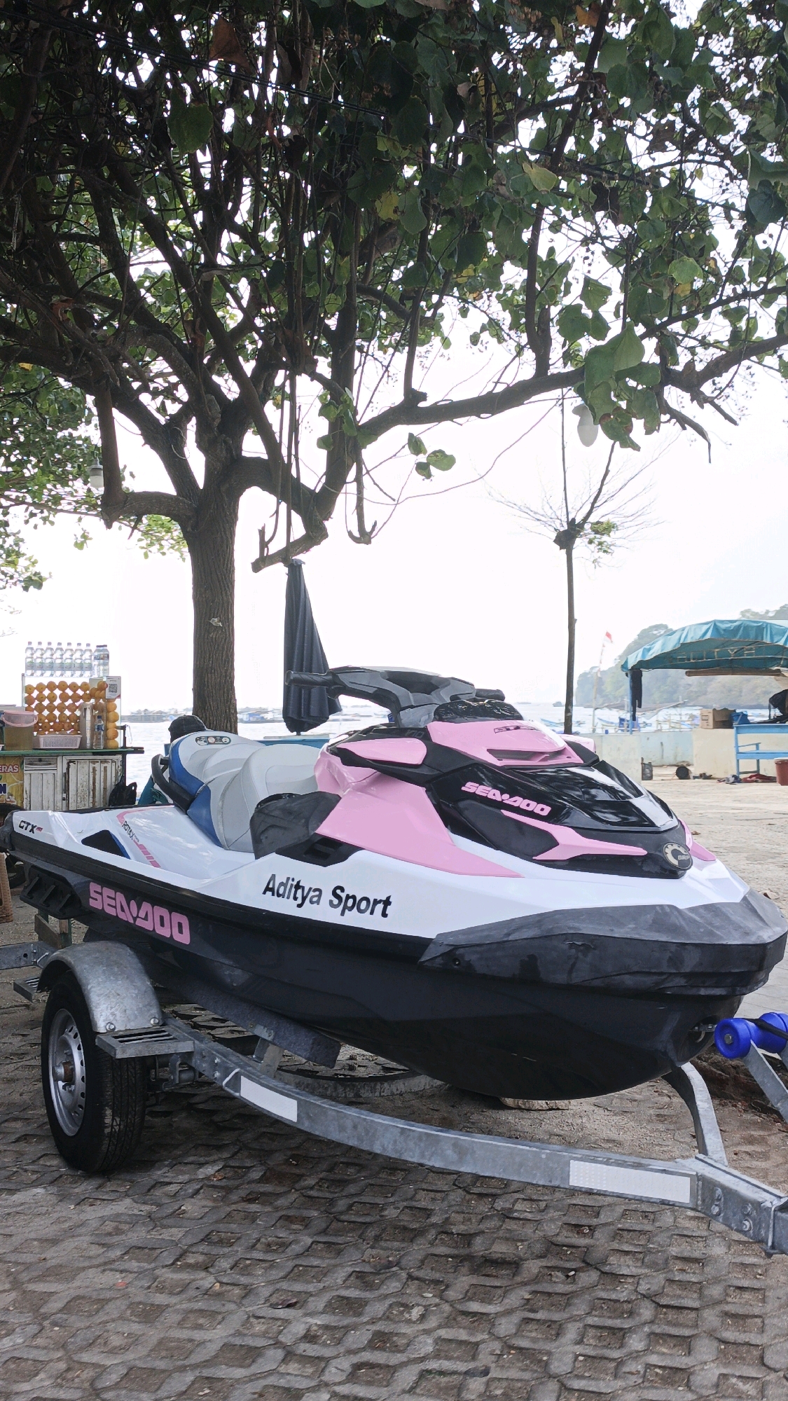 In Desa Pagandaran, Indonesia, a pink and white Sea-Doo jet ski, labeled "Aditya Sport," rests on a trailer beneath a large tree. The jet ski is parked on a paved surface, likely a beachfront promenade or parking area, with a sandy beach and the ocean visible in the background. Other elements of the scene include a refreshment stand with bottled water and oranges, and various beachgoers and structures further in the distance.