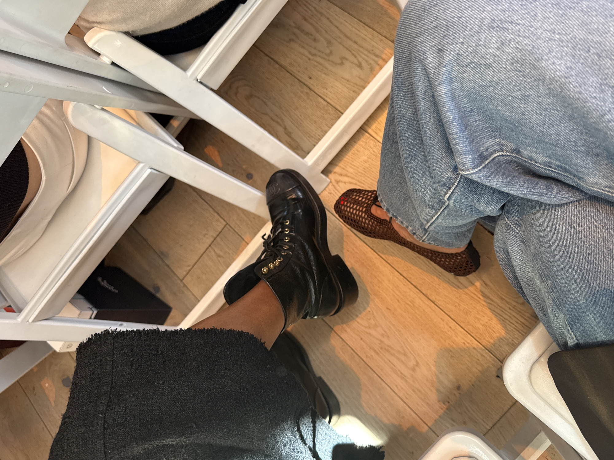 The image is a close-up, overhead shot of legs and feet in an indoor setting, likely within a building in the city of Los Angeles, United States of America. There are three people visible from the knees down.
One person is wearing a dark-colored, possibly black, textured skirt and black leather boots with gold eyelets. Another person is wearing loose-fitting, light-blue denim jeans and brown woven shoes. Part of a third person wearing a cream-colored garment is visible in the upper left corner.
The people are seated on white folding chairs on a light-colored wooden floor. A black box is partially visible under one of the chairs. The lighting appears to be indoor artificial lighting. There are no visible signs indicating a specific location or interaction.