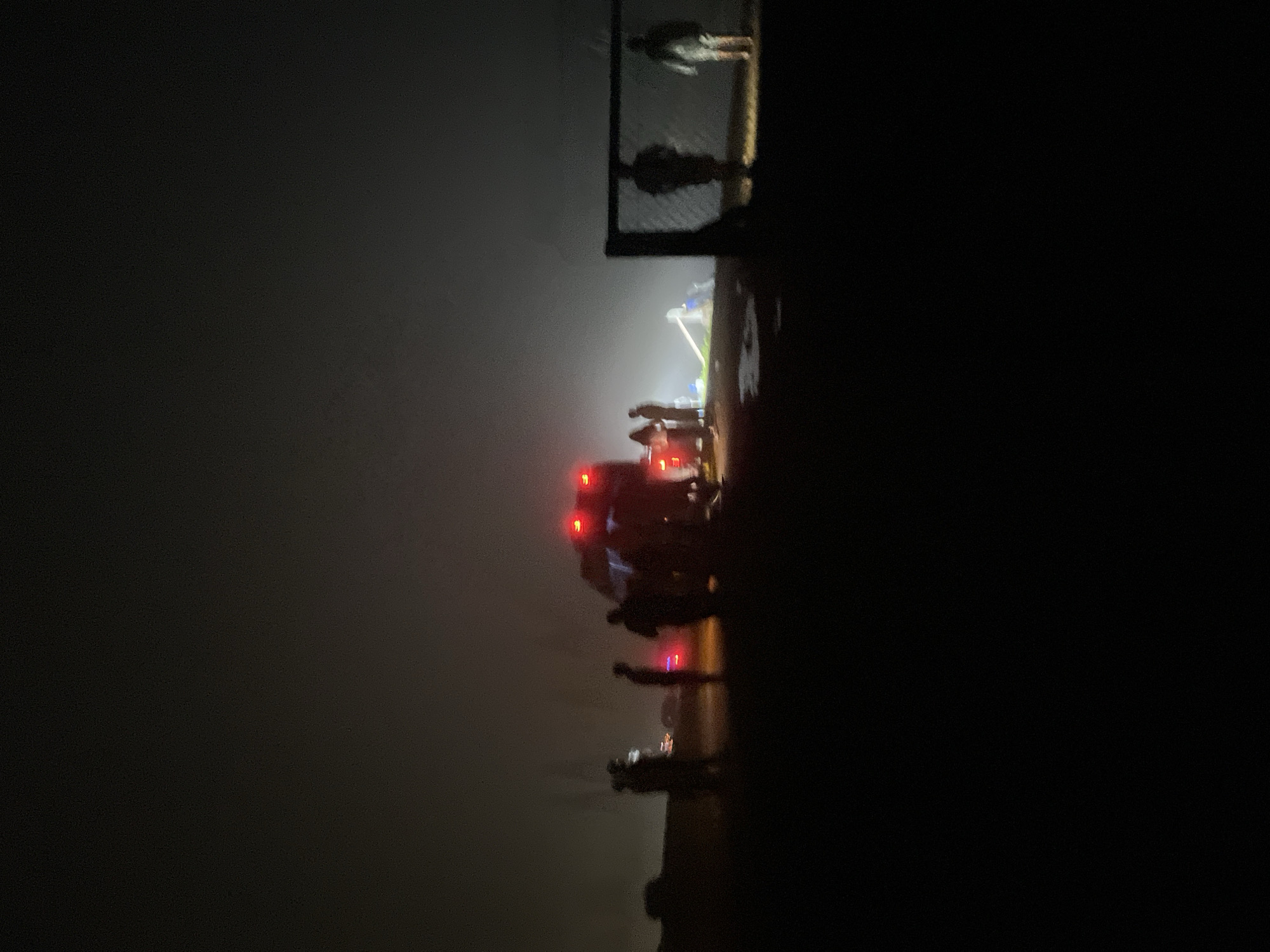 At night, in a foggy, dark environment, a group of people are gathered around an emergency vehicle, possibly an ambulance, with its red lights illuminated. The people are silhouetted against the dim light and fog, making their features indistinguishable. Some individuals appear to be interacting with each other or observing the scene. In the background, a fence-like structure is visible, with two silhouetted figures standing near it. The ground is dark and uneven. The scene suggests a potential emergency situation or a gathering responding to an event. The location is specified as Gomismta, Georgia.
