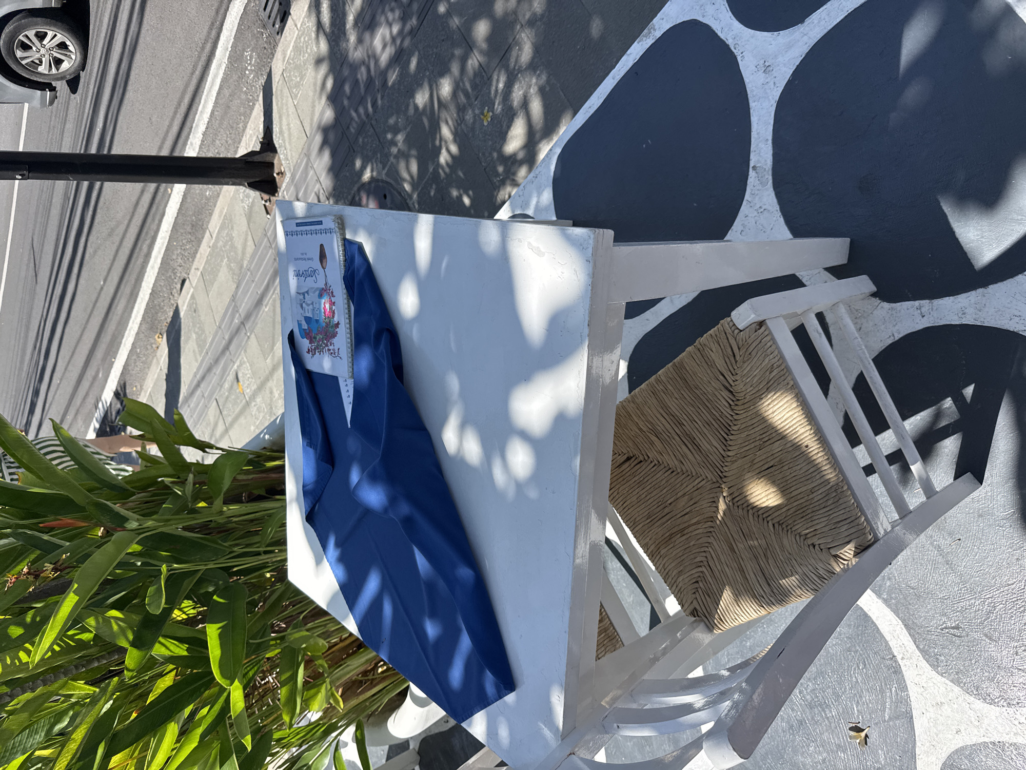 The scene is an outdoor setting in what appears to be Denpasar, Indonesia, indicated by the Greek-themed restaurant guide on the white table. A white wooden chair with a woven seat sits next to the table. A dark blue shirt lies on the table next to a notebook or guide for a Greek Restaurant in Santorini. A green plant with large leaves sits on the left corner of the table. The ground has a distinct white-and-black abstract pattern. A section of a road is visible in the background, along with a car tire and a street lamp.