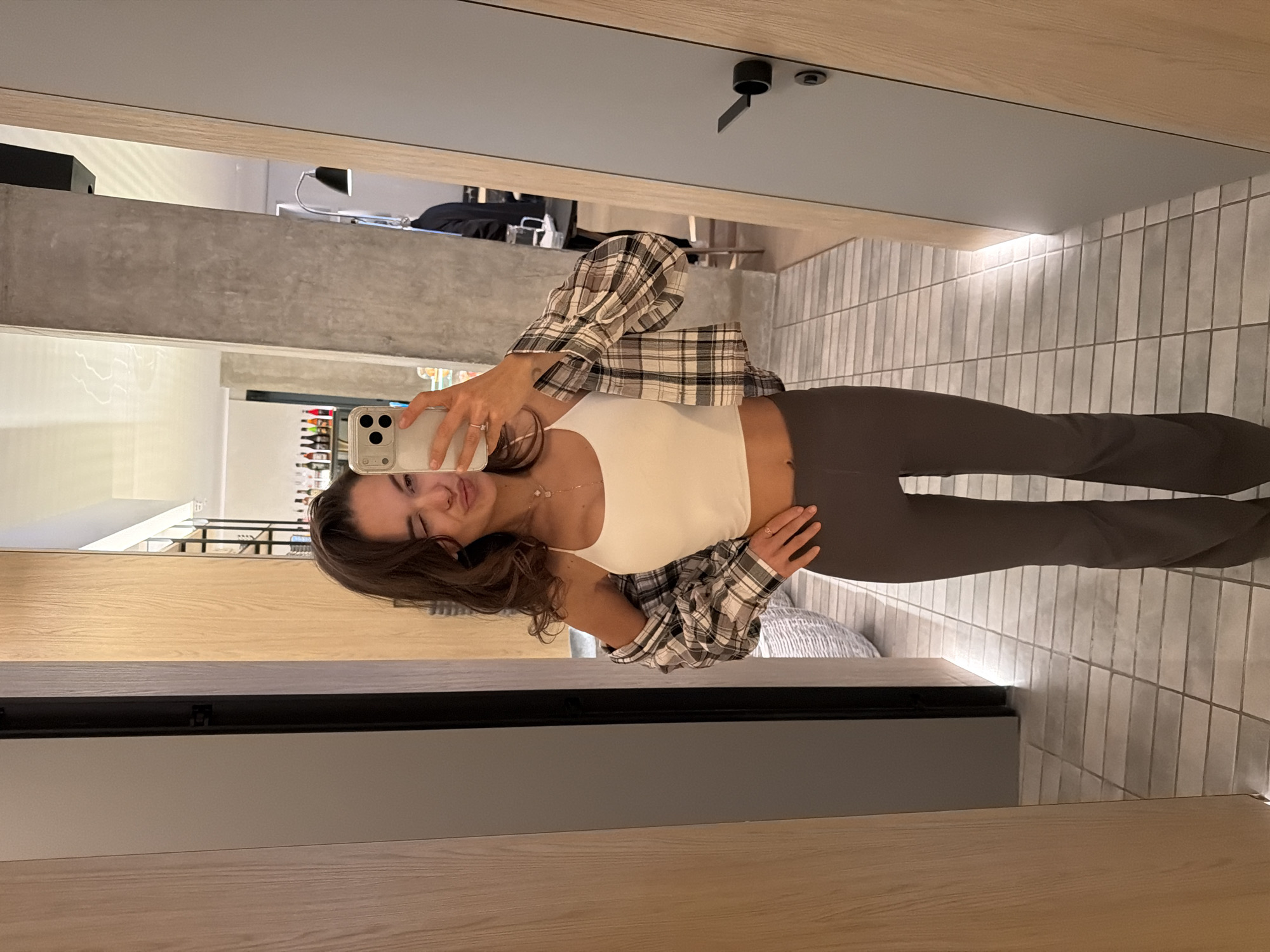 A young woman takes a selfie in a mirror. She is wearing dark leggings, a white tank top, and a plaid shirt. The background appears to be a room with tiled walls, possibly a dressing room or retail space. In the mirror's reflection, there is a glimpse of what might be a bar or storage area with various bottles. There is no information in the image to identify a location in Krasnoyarsk, Russia.