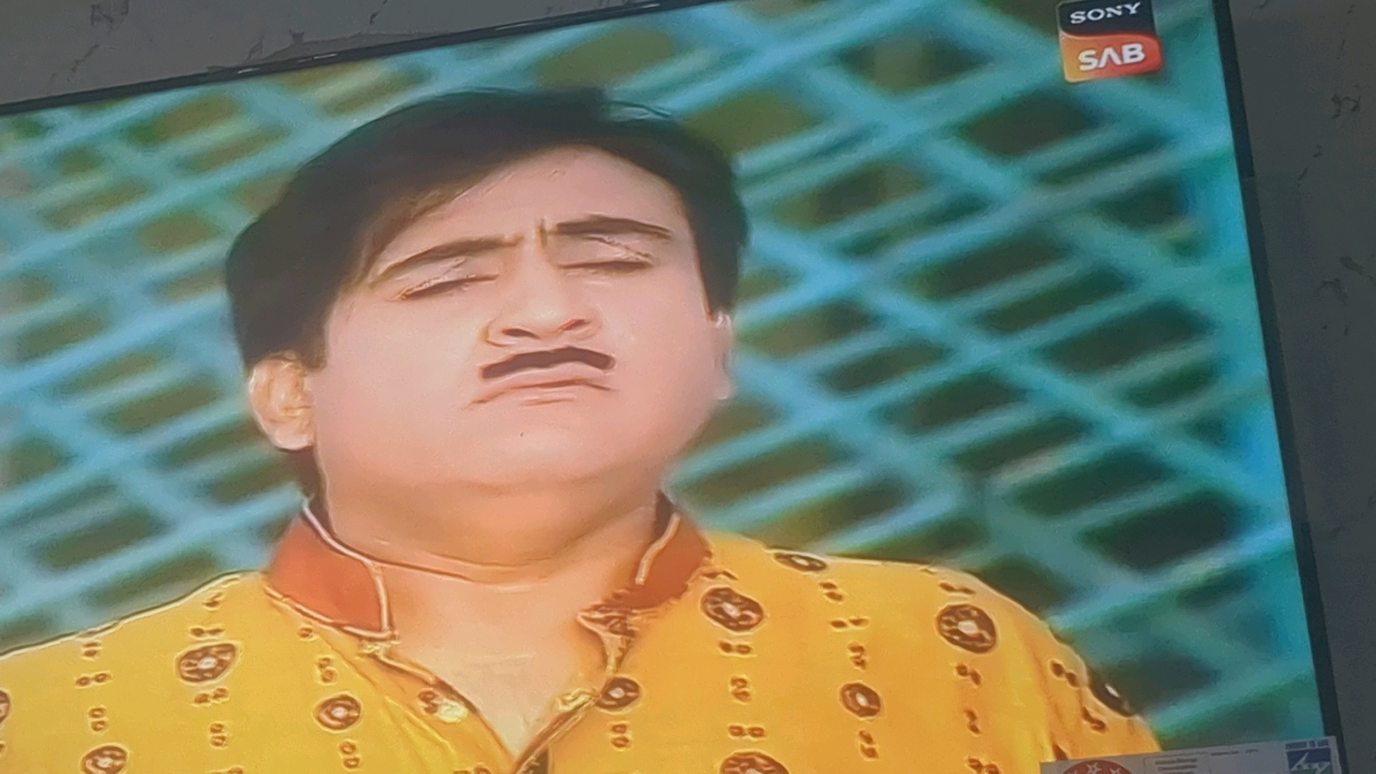 An Eastern Indian man with dark hair and a mustache is shown from the chest up, on a television screen. He is wearing a yellow shirt with a red collar and circular brown patterns. His eyes are closed and his face is contorted in an exaggerated expression, with his cheeks puffed out and his mouth wide open. The background of the screen is a blue and green lattice pattern. The Sony SAB logo is visible in the upper right corner of the screen. The image appears to be a still from a television show or movie.