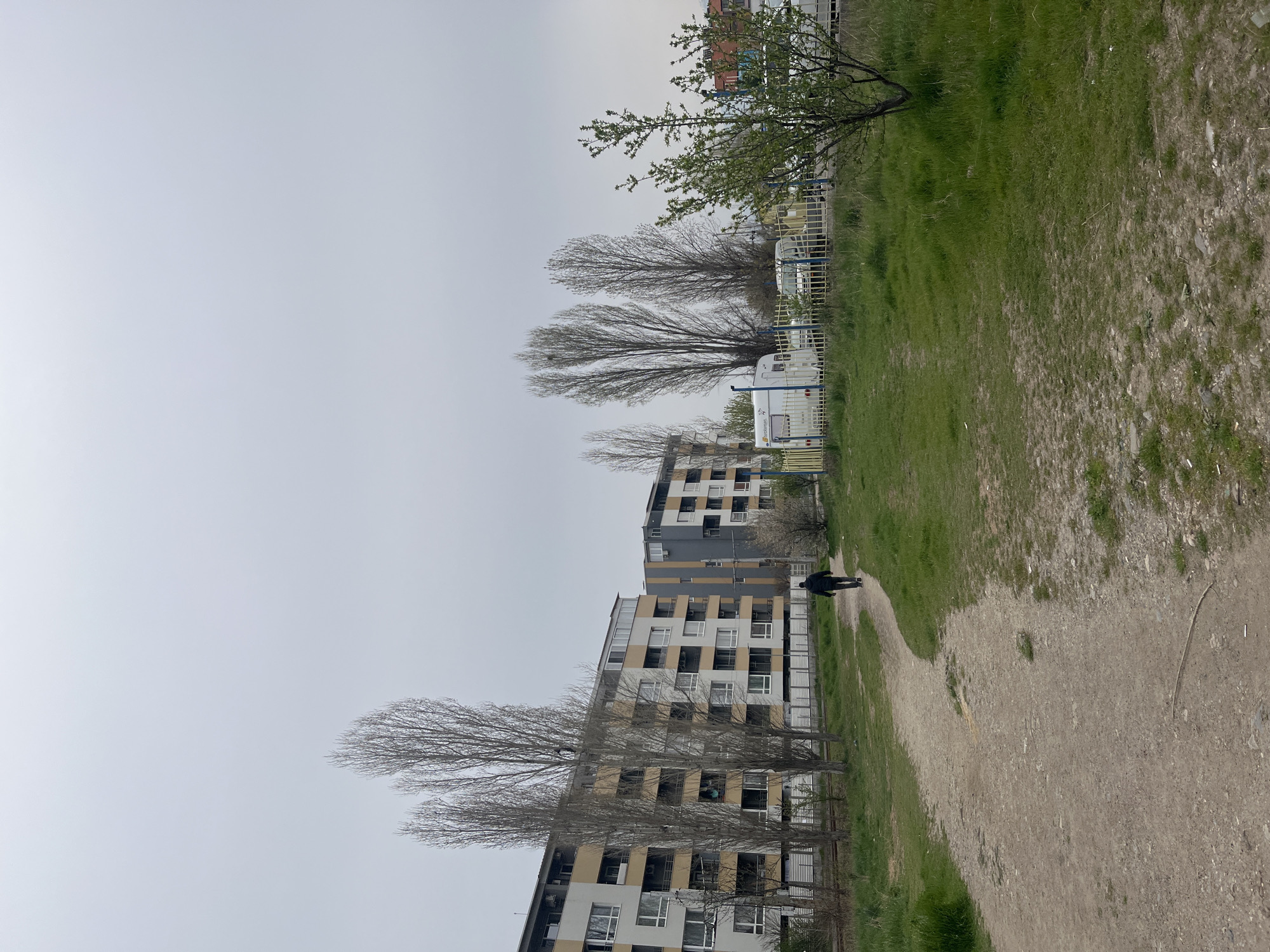 The image depicts an urban park or green space in Sofia, Bulgaria. Multiple apartment buildings are visible on the left side of the image with bare trees in front of them. A chainlink fence runs between the buildings and the green space. Several trailers are parked beyond the fence in the upper part of the image. A person is walking along a dirt path through the green space. The sky is overcast.