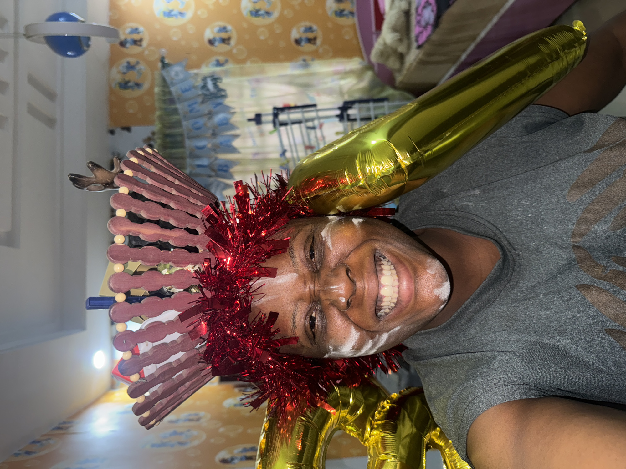 The image shows a person with white face paint, a red tinsel garland on their head, and a headdress made of painted craft sticks and small plastic reindeer antlers. A gold balloon in the shape of a number is partially visible. The person is smiling broadly. The background includes a patterned wall, diapers stacked in a rack, and a ceiling fan. The location is Unknown, Unknown.