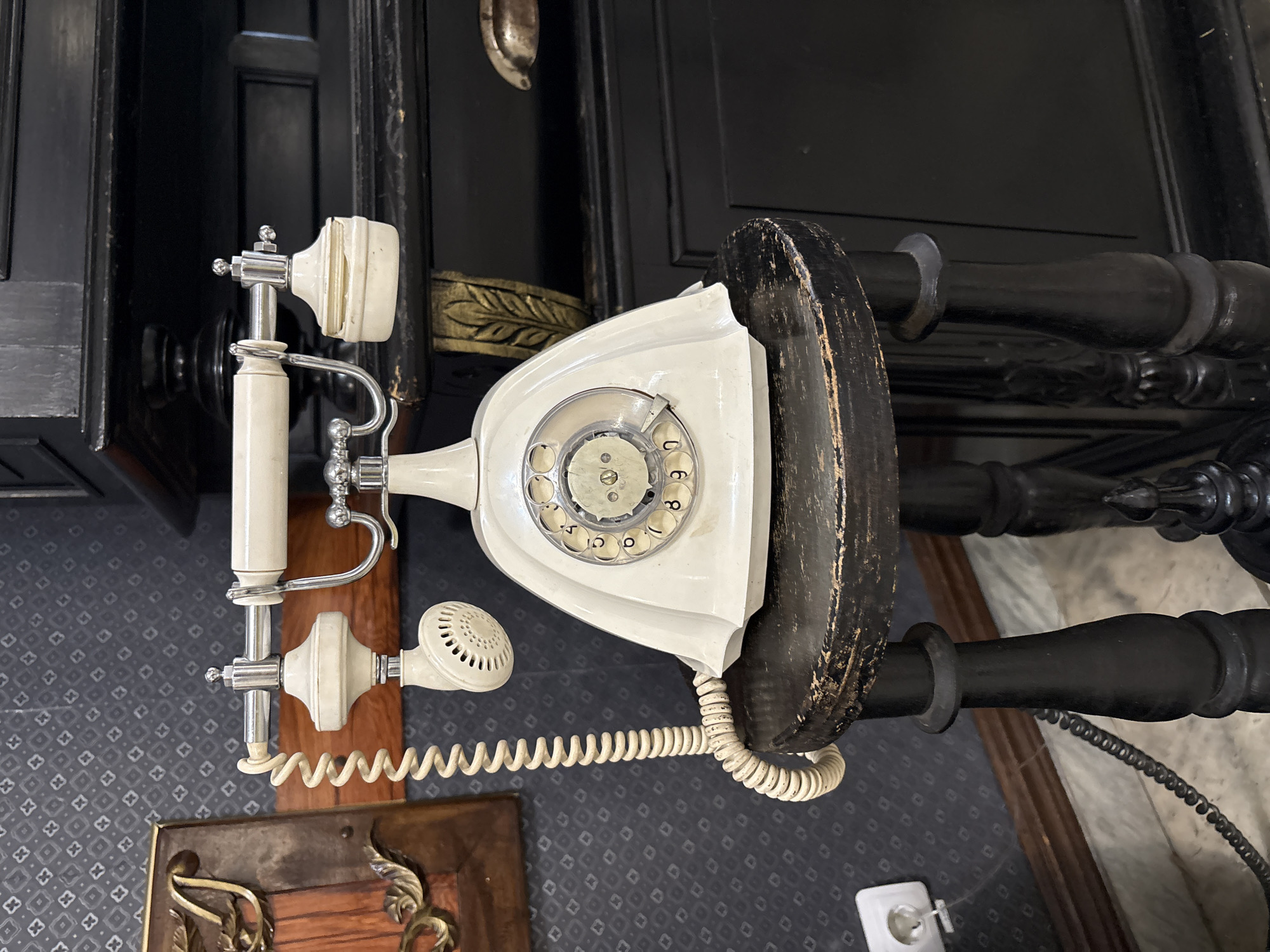 The image shows an antique white rotary dial telephone with a black wooden base and ornate black wooden legs. The telephone is positioned on a dark, patterned carpet. The handset is separate from the base, attached by a coiled white cord. A similar coiled cord connects the handset to the receiver. The dial has numbers from 0 to 9. The black wooden furniture behind the telephone appears to be a cabinet or table with decorative carvings and a metallic leaf-like accent. The scene is indoors, and the lighting suggests it could be daytime. There is no visible text in the image. The overall impression is of a vintage setting, possibly a home or an antique shop in Zavod Mikhailovskii, Russia.