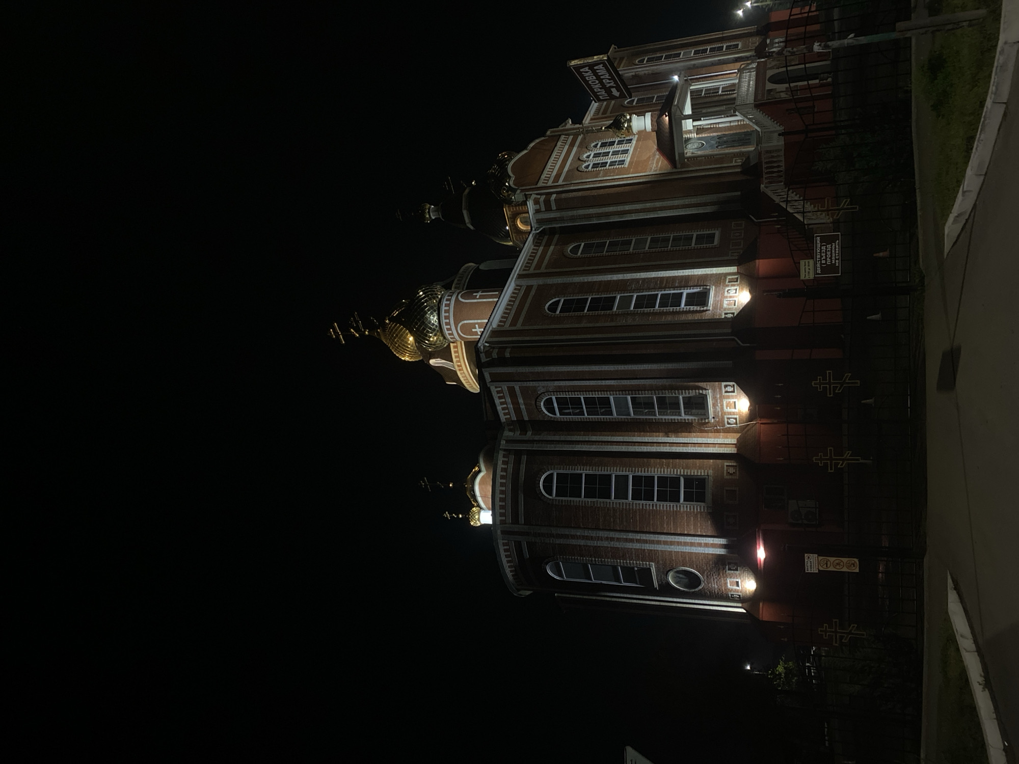 The image depicts a multi-story brick building with ornate, golden domes, likely a church, illuminated against a dark night sky. The building's architecture includes arched windows and decorative trim. A fence runs along the building's perimeter on the right side of the image, with a paved area beside it. A sign with lettering in Cyrillic is attached to the building. The location is Unknown, Unknown.
