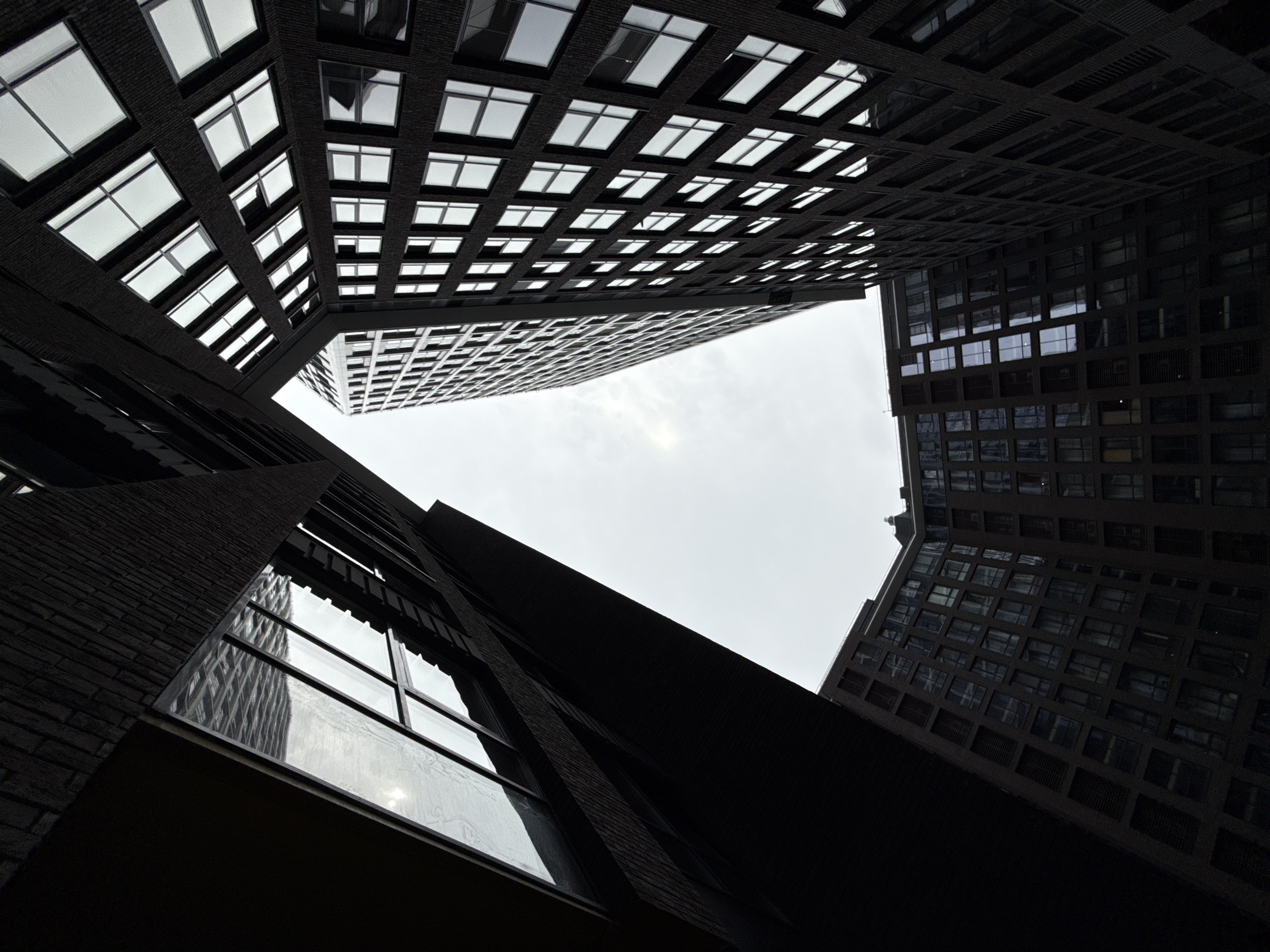 The image shows a low-angle view looking up at modern, dark-colored apartment buildings that create a central void of overcast sky. The buildings feature numerous windows with a grid-like pattern, reflecting the sky and some of the building's structures. On the left, a building with a darker, more textured facade, possibly brick, is visible, with a large, angled window reflecting a patterned surface. To the right and in the background, taller buildings with a more uniform facade and densely packed windows rise up. The architecture is contemporary, with sharp lines and geometric forms. The overall impression is one of urban density and imposing architecture, with a stark contrast between the dark buildings and the bright, cloudy sky. The style of the architecture, particularly the facade treatment and window design, is characteristic of contemporary residential complexes found in Moscow, Russia.