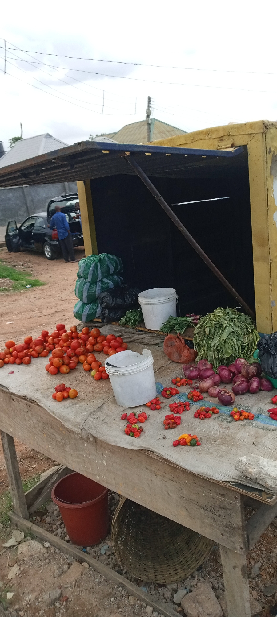 The image depicts a roadside vegetable stall in Jos, Nigeria. A wooden table displays tomatoes, Scotch bonnet peppers, red onions, and leafy greens. Two white buckets and a red bucket are visible near the produce. Three green striped bags are stacked behind the table, near a black car with the driver's side door and trunk open. A man stands near the car. The stall has a partially open, yellow corrugated metal structure. The ground is dirt. Overhead are utility wires and the roofs of nearby buildings.