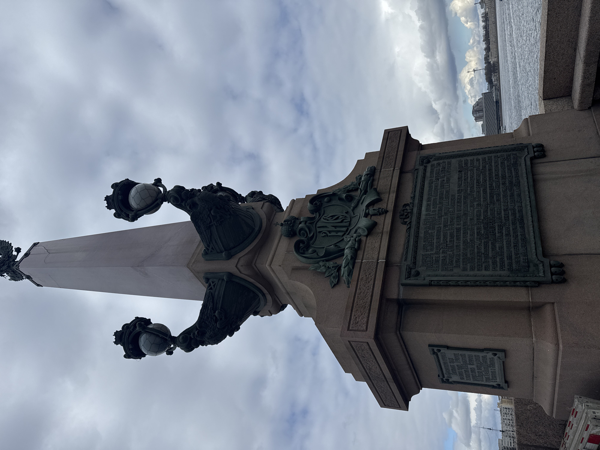 The image shows a detailed view of a monument or lamppost in Saint Petersburg, Russia. The monument features an obelisk shape and ornate bronze or ironwork decorations, including lamps with globe-shaped fixtures. There are also plaques with Cyrillic inscriptions on the base of the monument. In the background, the Neva River can be seen, along with buildings lining the embankment under a cloudy sky. The monument is likely part of a bridge, given the river view and the architectural style.