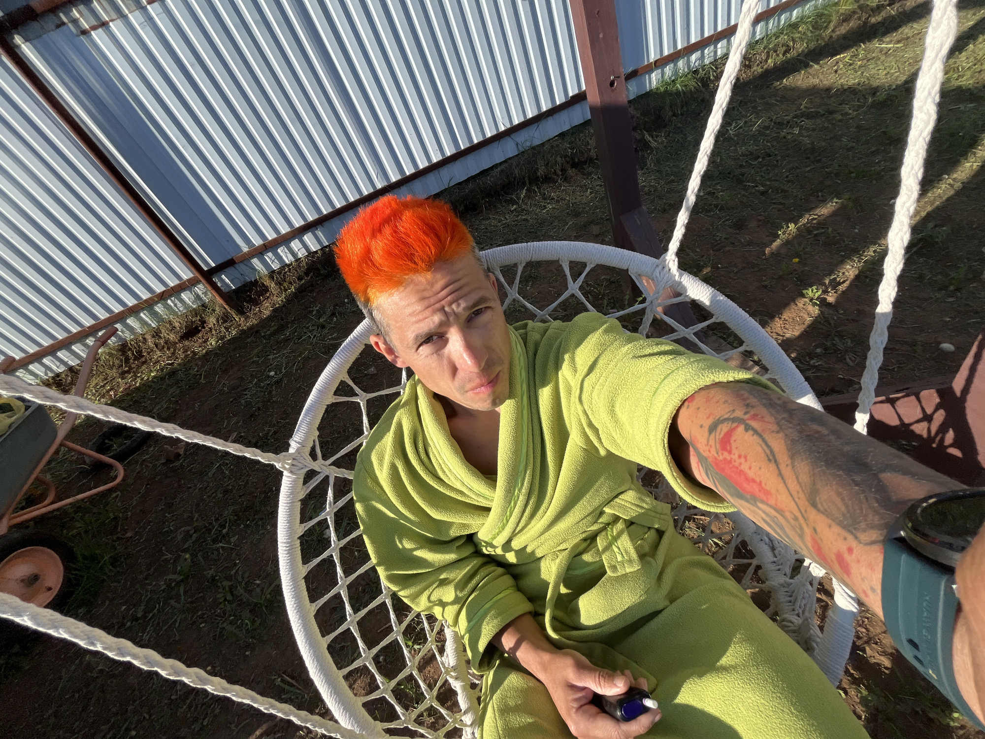 The photograph depicts a man with bright orange hair, wearing a green bathrobe, sitting in a white macrame swing chair. The setting appears to be outdoors, with a corrugated metal roof structure visible in the background. The ground shows grass and dirt. The man has tattoos on his arm, and a blue object in his hand. He is also wearing a green watch. The photograph is taken from a high-angle perspective. There is no specific visual evidence within the image to confirm that the location is Chaikovsky, Russia.
