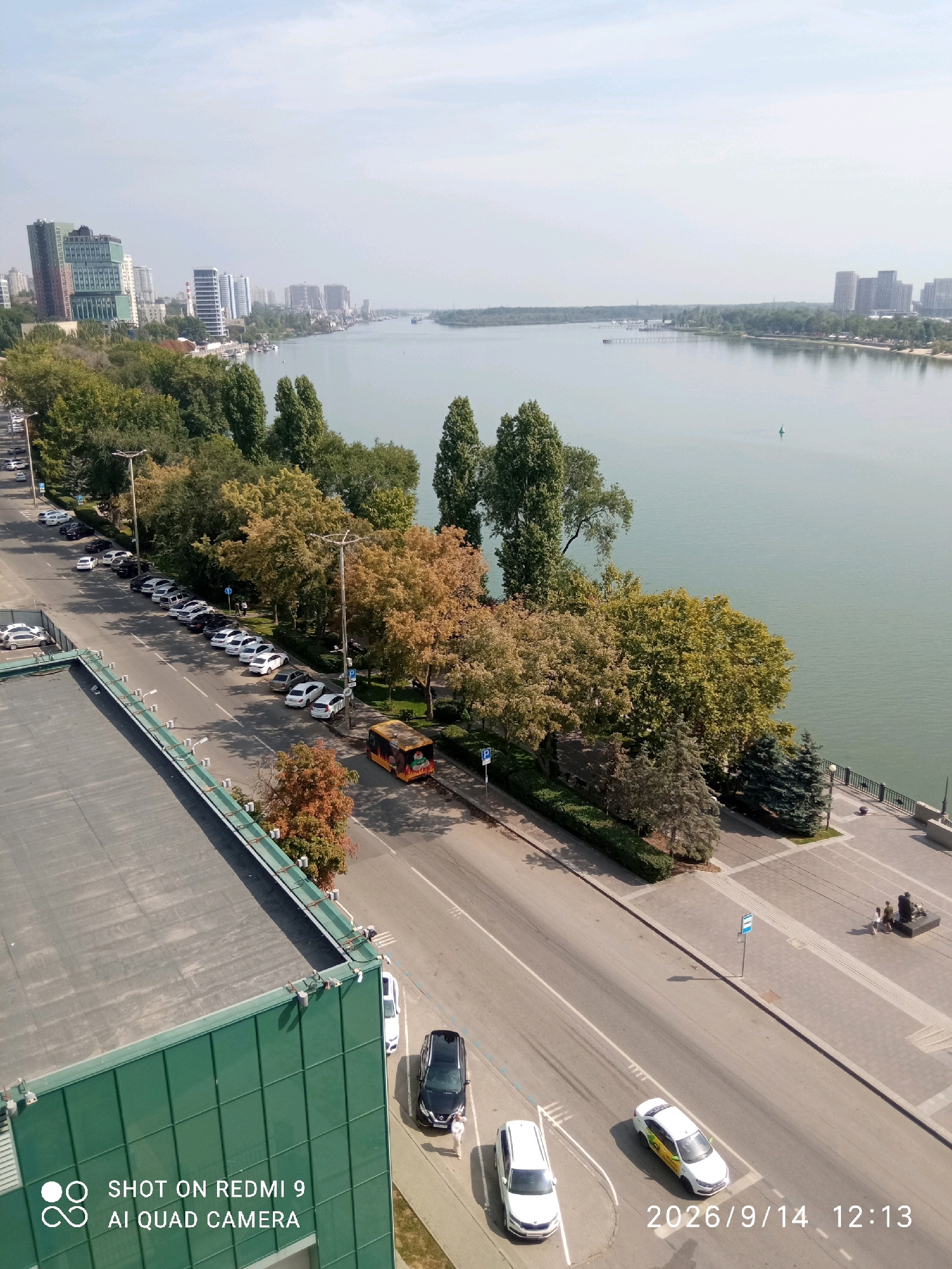 The aerial image captures a panoramic view of the Don River embankment in Rostov-on-Don, Russia. On the left, a multi-story building with a green roof is visible, adjacent to a street lined with parked cars. The street runs parallel to a promenade bordering the river. A yellow bus with an advertisement on its side is parked on the right side of the street. The promenade features trees with autumn foliage, benches, and a few people. The Don River stretches across the frame, with a distant cityscape on the opposite bank. The sky is hazy and overcast. The timestamp "2026/9/14 12:13" is overlaid in the lower right corner.