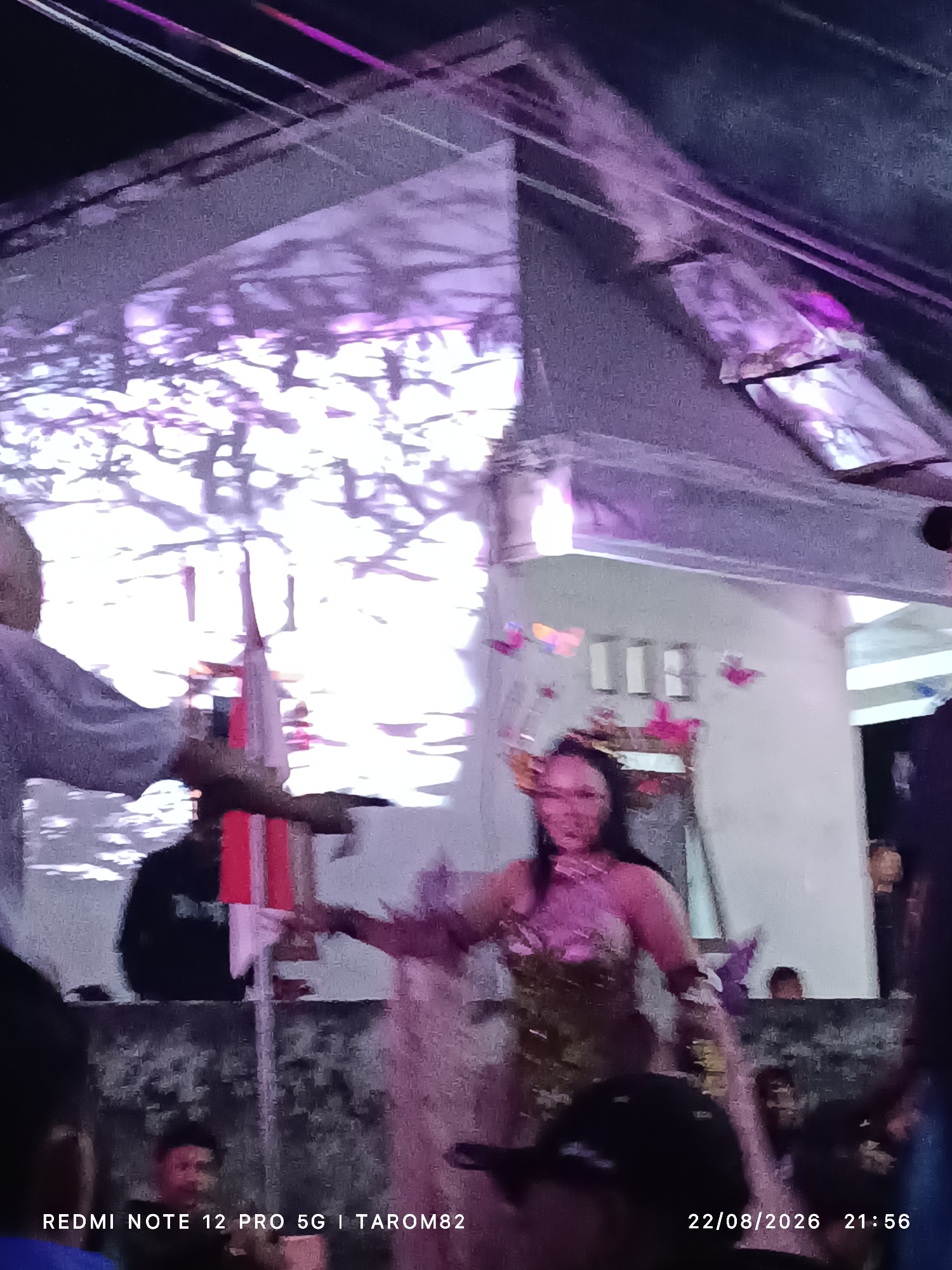 This is a low-resolution, dimly lit photograph taken outdoors at night in Mundusewu, Indonesia. The scene appears to be a gathering or event with a performance. In the foreground, several blurry figures are visible, including a person in what seems to be a costume with a pink top and a patterned lower garment. This person is the central focus and is looking towards the camera. Behind them, a white pole with red and white fabric wrapped around it stands upright, possibly part of a decoration or prop. The background is an indistinct mix of structures and the sky, with purple and pink lighting casting shadows and highlights. The lighting suggests stage or event illumination. A timestamp at the bottom of the image reads "22/08/2026 21:56" and "REDMI NOTE 12 PRO 5G | TAROM82".