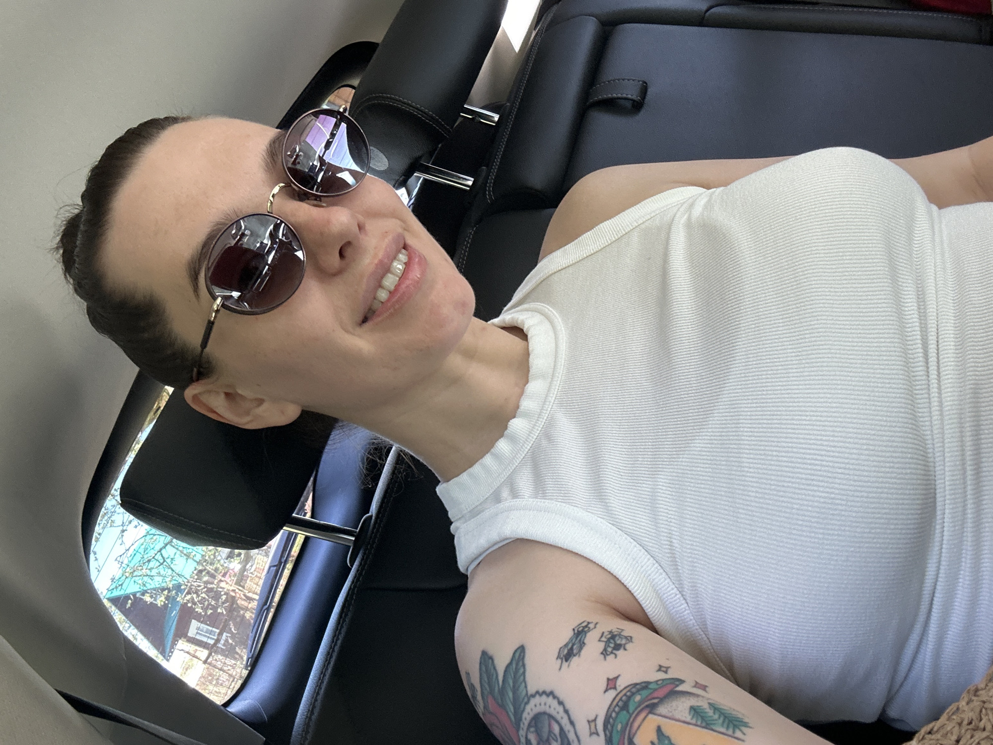 A woman is reclining in the back seat of a car, angled towards the camera. She is wearing round sunglasses with a brown tint and a white ribbed tank top. Her hair is pulled back. Part of a tattoo is visible on her upper arm. Outside the car window, trees and a glimpse of a building are visible. The interior of the car has black leather seats and headrests. The location information is not discernible from the image.