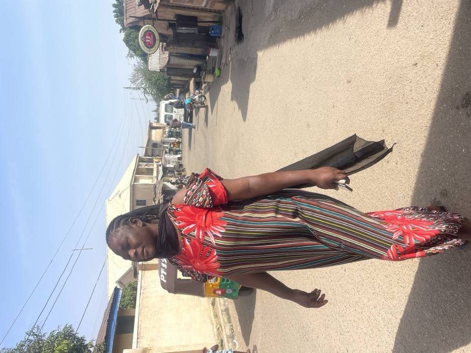 This image captures a daylight street scene in Jos, Nigeria. The primary subject is an adult woman of African descent, shown mid-stride walking from the right foreground towards the left. She is wearing a long, loose-fitting dress featuring a red background with large red floral patterns on the chest and lower portion, interspersed with vertical stripes of various colors including red, green, yellow, black, white, and blue. The dress has cold-shoulder sleeves with ruffle details. A black fabric scarf is draped around her neck and over her right shoulder, with a long end hanging down. Her braided hair is pulled back, and her gaze is directed slightly upwards and to the left with a neutral expression. She holds a small, light-colored object in her right hand.
The street is paved with a light brown asphalt surface. Strong sunlight illuminates the scene, casting distinct shadows towards the right side of the frame.
In the midground on the left, a light-colored wall is visible, behind which stands a building with a dark roof. A green and brown auto-rickshaw (keke Napep), marked with "PT-19," is parked on the roadside. Further down, a multi-story building with a light-colored facade and roof is present. Overhead power lines are visible against the clear blue sky.
On the right side of the street, a sequence of compact, informal structures with corrugated metal roofs and walls, likely commercial stalls, line the road. A prominent circular sign displaying "33" and additional text, characteristic of a beverage advertisement, hangs from one of these structures. Further along the street, a white van, a motorcycle, and several individuals are discernible. Various containers, including buckets and jerrycans, are placed on the ground next to the structures.