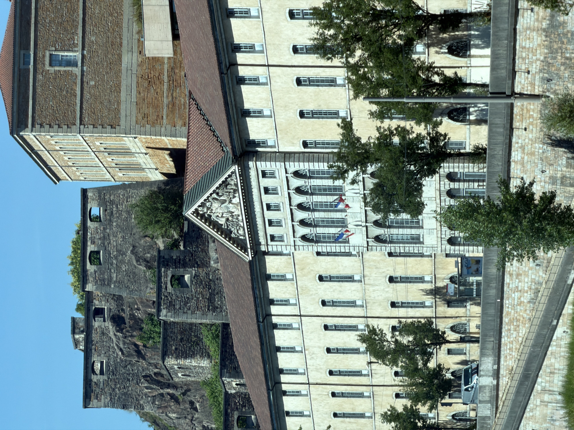 The image depicts the Fort Saint-Jean in Lyon, France, a historic military fortification. The fort is characterized by its stone construction, with visible ramparts, battlements, and arched windows. The architecture suggests a historical period of military defense, with rough-hewn stone juxtaposed against more refined brickwork on a larger building in the foreground.
In the foreground, a large, multi-story building with a pale facade and a red-tiled roof stretches across the image. This building features numerous windows, many of which are tall and narrow, with a rhythmic repetition that suggests a formal architectural style. A decorative pediment with sculpted figures is visible at the peak of the building's facade on the left. French flags are displayed on the facade of this building.
To the left and behind the foreground building, the older stone fortifications of Fort Saint-Jean are visible, built into a rocky hillside. These fortifications are more rugged and appear older, with evident stonework and openings that suggest defensive emplacements. A separate, taller brick building with a peaked roof is visible to the right of the stone fort, showcasing a different architectural style.
At the base of the buildings, a road is visible, with a white van driving on it. A concrete barrier separates the road from a lower level, which appears to be a walkway or viewing area. This lower level is constructed with stone blocks. Beyond this, a sloped embankment covered in grass suggests the proximity of a river or natural landscape. Trees are scattered throughout the scene, some appearing in front of the buildings and others on the fortifications. The sky is clear and blue, indicating fair weather.