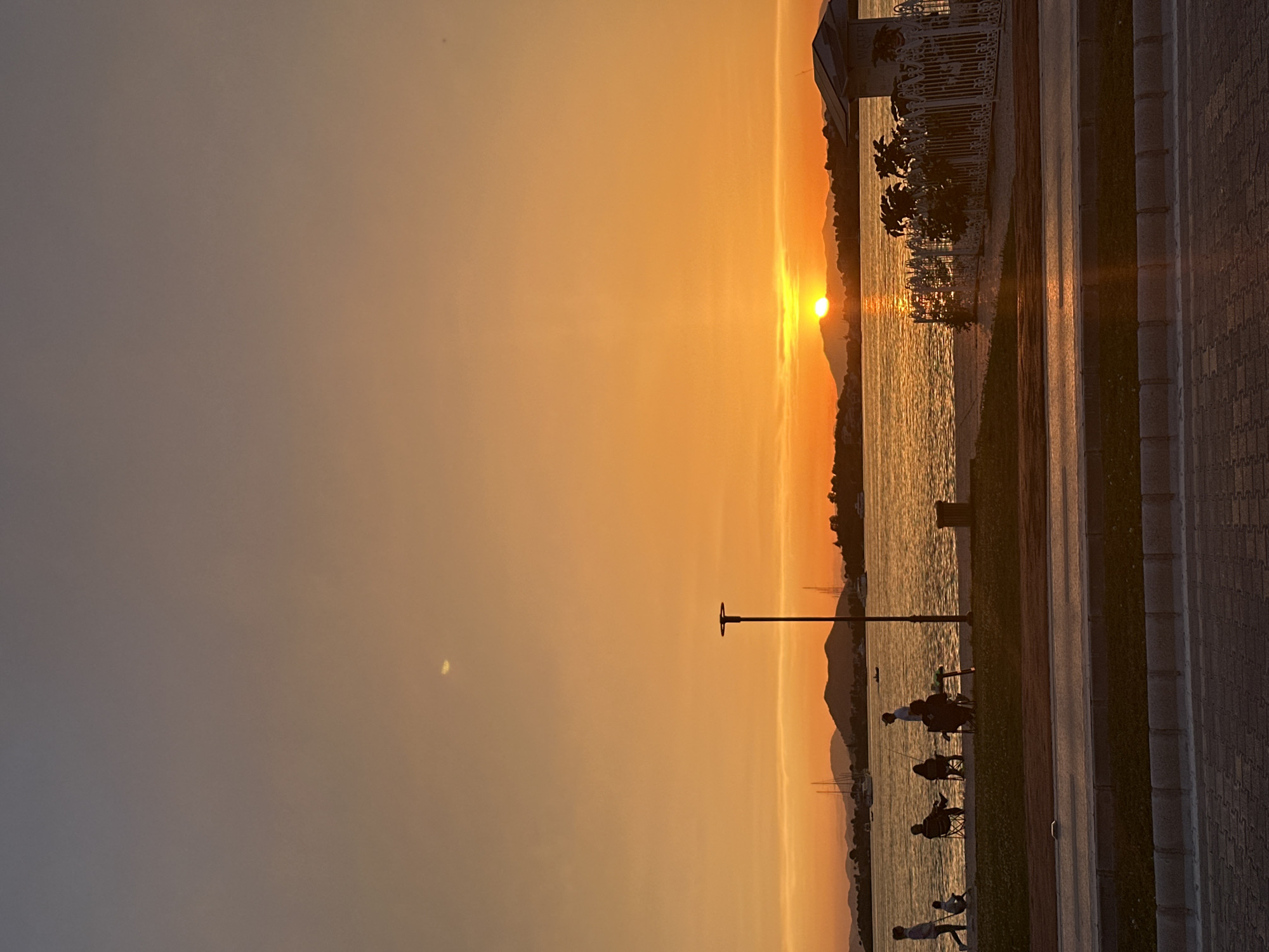Captured in Fethiye, Turkey, the image depicts a serene sunset setting over the water. Silhouetted figures are seen along the shore, likely residents or visitors enjoying the evening. They appear to be fishing or simply watching the sun descend. A lamppost stands nearby, and in the distance, mountains or hills are faintly visible. The sky exhibits a gradient of orange hues, reflecting on the water's surface. A paved walkway with some vegetation lines the edge of the shore. The overall impression is that of a peaceful evening in a popular coastal city.