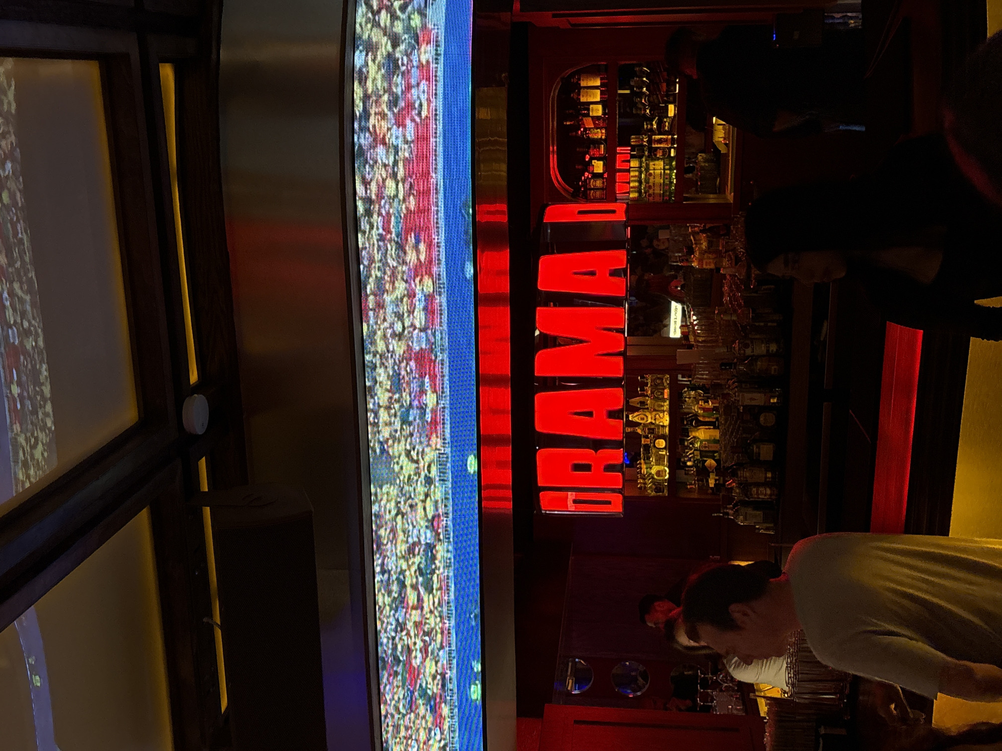 In San Pedro Garza García, Mexico, a bar's interior is depicted, featuring a large LED screen above a bar displaying a crowd in a stadium. The bar itself is illuminated with red neon lights that spell out "DRAMA," a prominent sign. Shelves behind the bar are stocked with various bottles of alcohol. Several individuals are present; one man in the foreground faces away from the camera, wearing a light-colored t-shirt, while a woman is partially visible in the foreground on the right, silhouetted against the bar. Other patrons are present but less distinct in the background. The environment is dimly lit, with red and yellow lighting contributing to a vibrant atmosphere.