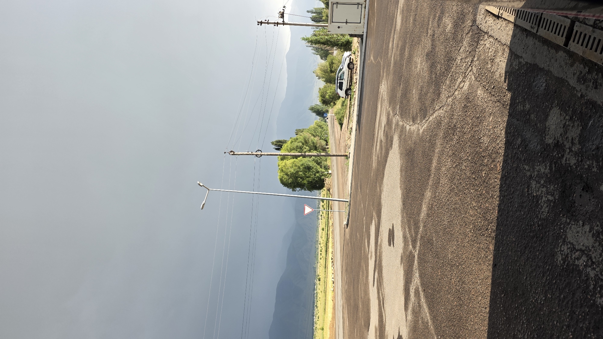The image captures a scene at an intersection in Kochkor, Kyrgyzstan, under a dramatic sky. A dark, overcast sky dominates the upper portion of the frame, with a sliver of lighter sky visible on the right. In the foreground, a wet asphalt road displays reflections and cracks, suggesting recent rain. A triangular yield sign is prominently positioned to the left of a tall, modern streetlamp. To the right, a weathered wooden utility pole with wires stretches across the frame, accompanied by a white metal enclosure. Beyond the road, a flat landscape unfolds with green fields and distant, hazy mountains. Several trees, some with full green foliage, are scattered across the mid-ground. A white car is parked on the side of the road near the utility pole. The overall impression is one of a rural or semi-rural environment on the outskirts of a town, with a sense of impending weather.