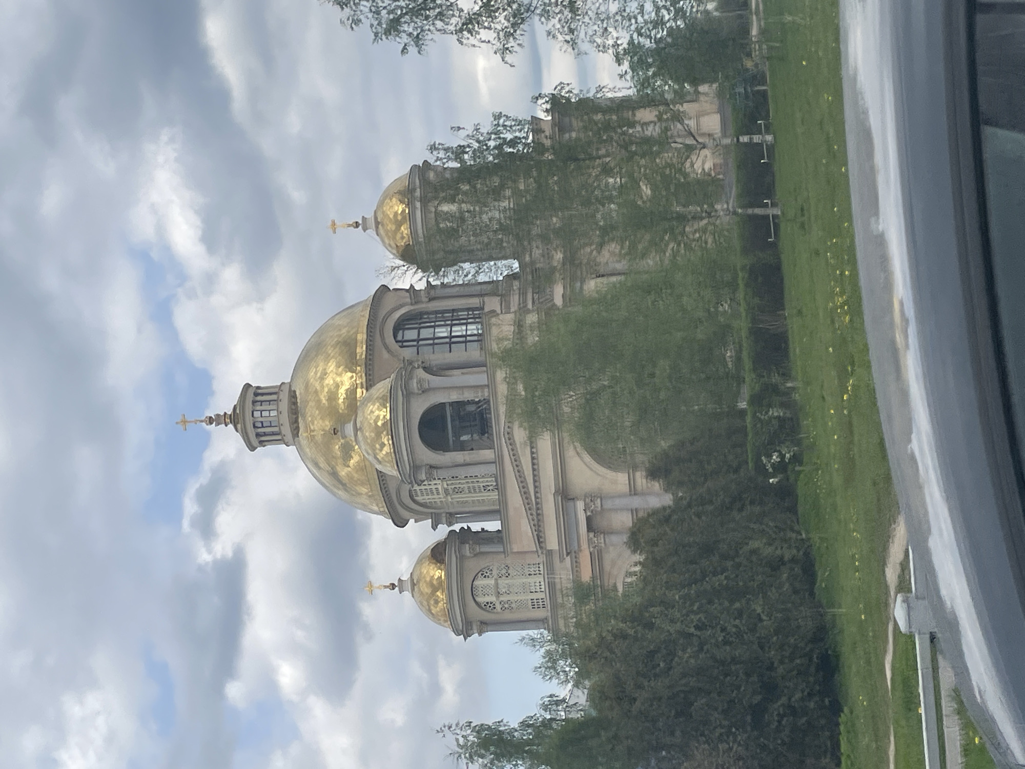 The image shows the Saint Petersburg Mosque in Saint Petersburg, Russia. The mosque has a large golden dome and several smaller ones, all topped with crosses. The architecture is reminiscent of Orthodox churches. The building is surrounded by trees and grass. The photo was taken from inside a car, judging by the car door visible on the right side of the frame. The sky is cloudy.