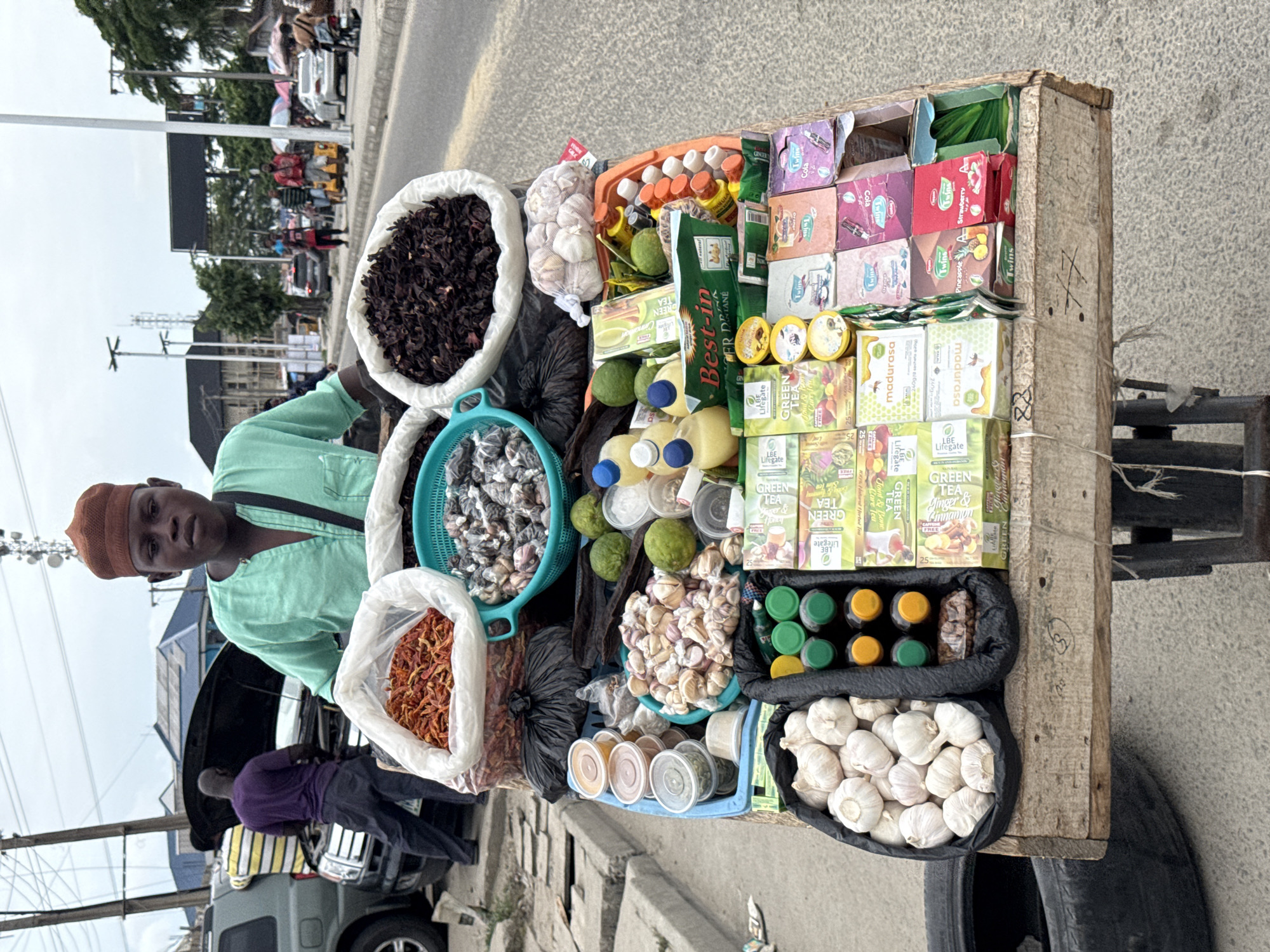 A street vendor in Ado, Nigeria, stands behind his cart, which is brimming with various goods for sale. He is wearing a brown skullcap and a light green shirt. The cart displays an assortment of items, including dried hibiscus leaves, spices, garlic, limes, and an array of packaged teas with brands such as "Lifegate" and "Twins." The vendor is looking at the viewer. In the background, a silver SUV with its hood open is visible, with people gathered around it, suggesting a possible car issue. The overall environment is that of a busy street scene.