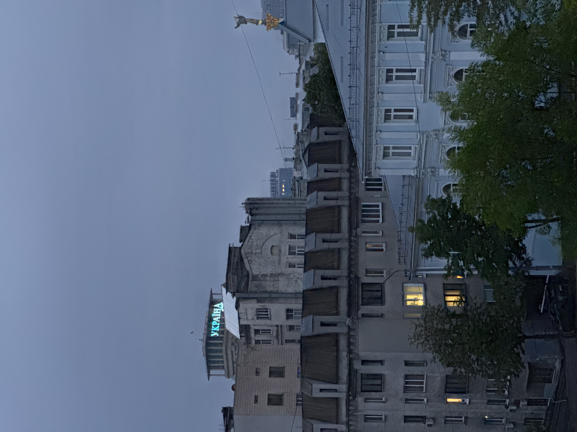 The image presents an urban landscape in Kyiv, Ukraine, captured during overcast conditions at dusk or early evening. The sky is a uniform, muted grey-blue. The scene is dominated by a dense collection of buildings of varying architectural styles and ages, interspersed with green trees.
In the foreground, a row of residential buildings features multiple windows, some illuminated with warm yellow light, suggesting occupancy. Air conditioning units are visible on several facades. Further right, a more ornate, light blue multi-story building stands out with decorative cornices and a mix of arched and rectangular windows. Trees with dense green foliage are scattered throughout, partially obscuring some structures.
Towards the mid-ground and left, another building prominently displays a glowing neon sign in Cyrillic script, reading "УКРАЇНА" (UKRAINA). In the far distance, atop a building roof, a golden statue of a female figure, likely a monument, is partially visible against the sky. Power lines crisscross the upper portion of the frame. No explicit human activity is visible, contributing to a quiet, somber atmosphere under the dim, natural light.