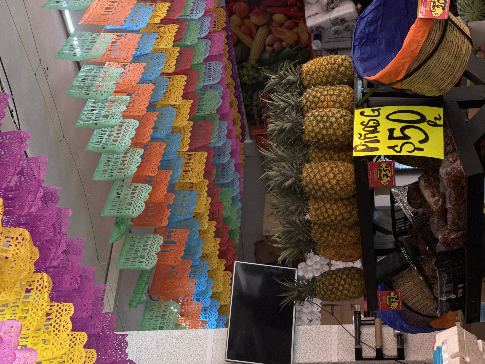 The image shows a grocery store in Puebla, Mexico, recognizable by the colorful papel picado (perforated paper) decorations hanging from the ceiling. A stack of pineapples is displayed for sale, with a sign reading "Piñas G $50 p/z" (Pineapples G $50 per piece). There are also items such as wrapped meat and other produce visible in the background. A black screen appears to be a part of the display. The environment seems like a busy market setting, with goods stacked high and a variety of items available.