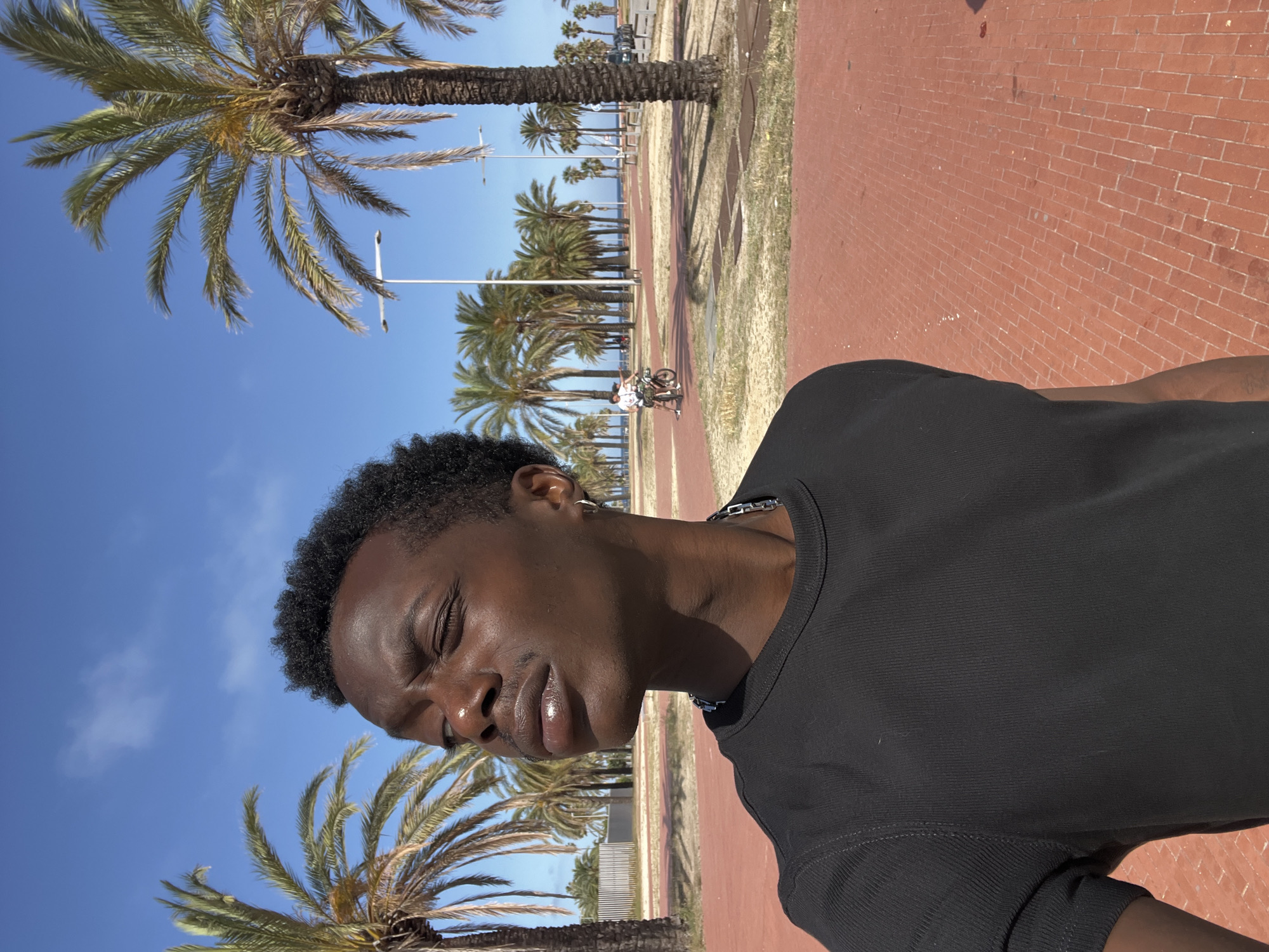 A young man with dark skin and curly hair is captured in a close-up shot, with his eyes closed and head tilted slightly upwards, suggesting a moment of relaxation or contemplation. He is wearing a black, ribbed t-shirt and a silver chain necklace. The background is a sun-drenched scene with palm trees lining a red brick pathway. In the distance, a person is seen riding a bicycle on the path. The sky is a clear, vibrant blue, indicating good weather, likely during the daytime given the strong sunlight casting shadows. The presence of palm trees and the distinct red brick paving strongly suggest a coastal promenade or park. The overall atmosphere is one of leisure and enjoyment, perhaps a vacation or a day out in a warm climate.