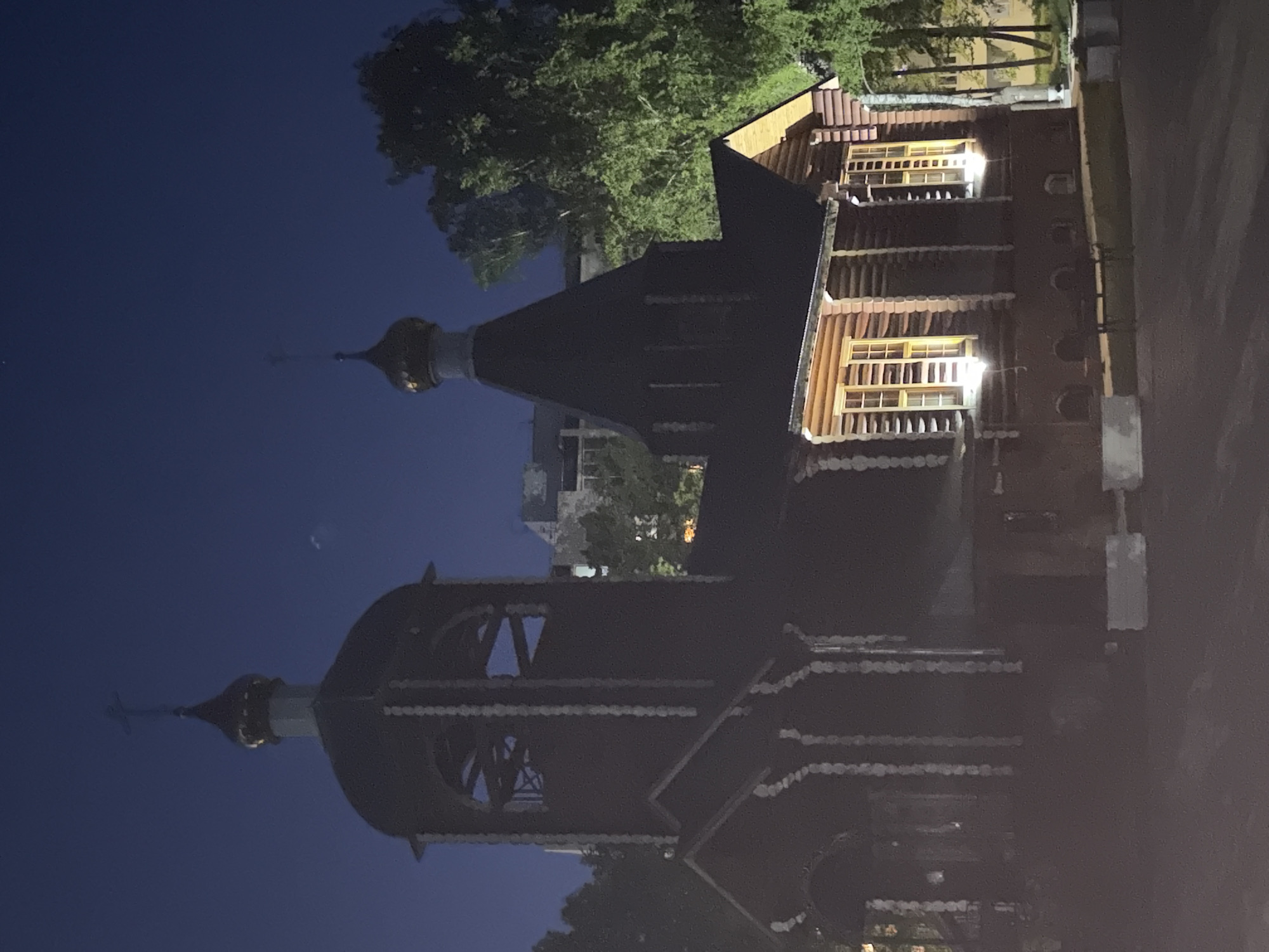 At night, the image shows a Russian Orthodox church and a log cabin building, possibly a monastery guesthouse, in Chaikovsky, Russia. The church has a dark dome with a golden cross, and the guesthouse has illuminated windows. The sky is a dark blue. Trees are visible in the background. The image appears to be rotated.