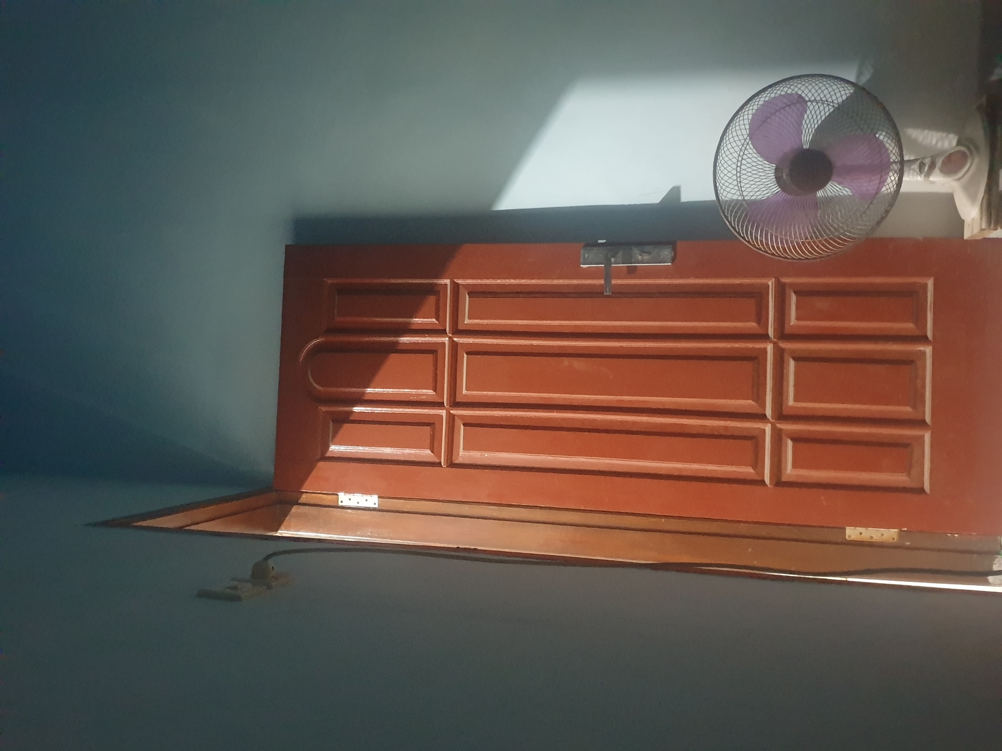 The image depicts an indoor scene, likely a room in Keboharan, Indonesia, featuring a reddish-brown wooden door ajar, revealing a sliver of light and a dark interior. The door has a paneled design and a metal door handle. To the right of the door, a white oscillating fan with purple blades is positioned on a surface, angled towards the right. The walls are painted a light blue color, with a distinct rectangular shadow cast across the wall from an unseen light source, likely sunlight, illuminating a portion of the wall and the fan. On the wall to the left of the door, a light-colored electrical outlet is visible with a black cable extending downwards. The lighting in the scene is dramatic, with strong contrasts between illuminated areas and shadows.