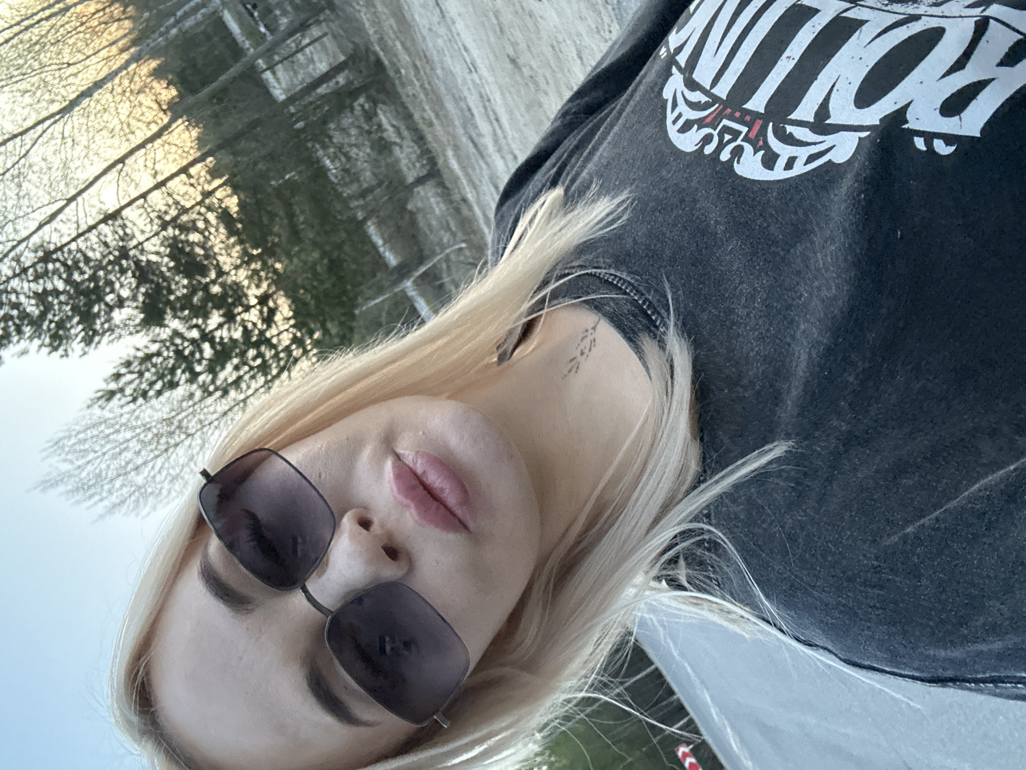 A woman with blonde hair, wearing sunglasses and a dark graphic t-shirt, takes a selfie. Her face is angled towards the camera. A small tattoo is visible on her neck. The background appears to be a winter scene with bare trees and snow, possibly in a wooded area. The location is Высокий, Russia.