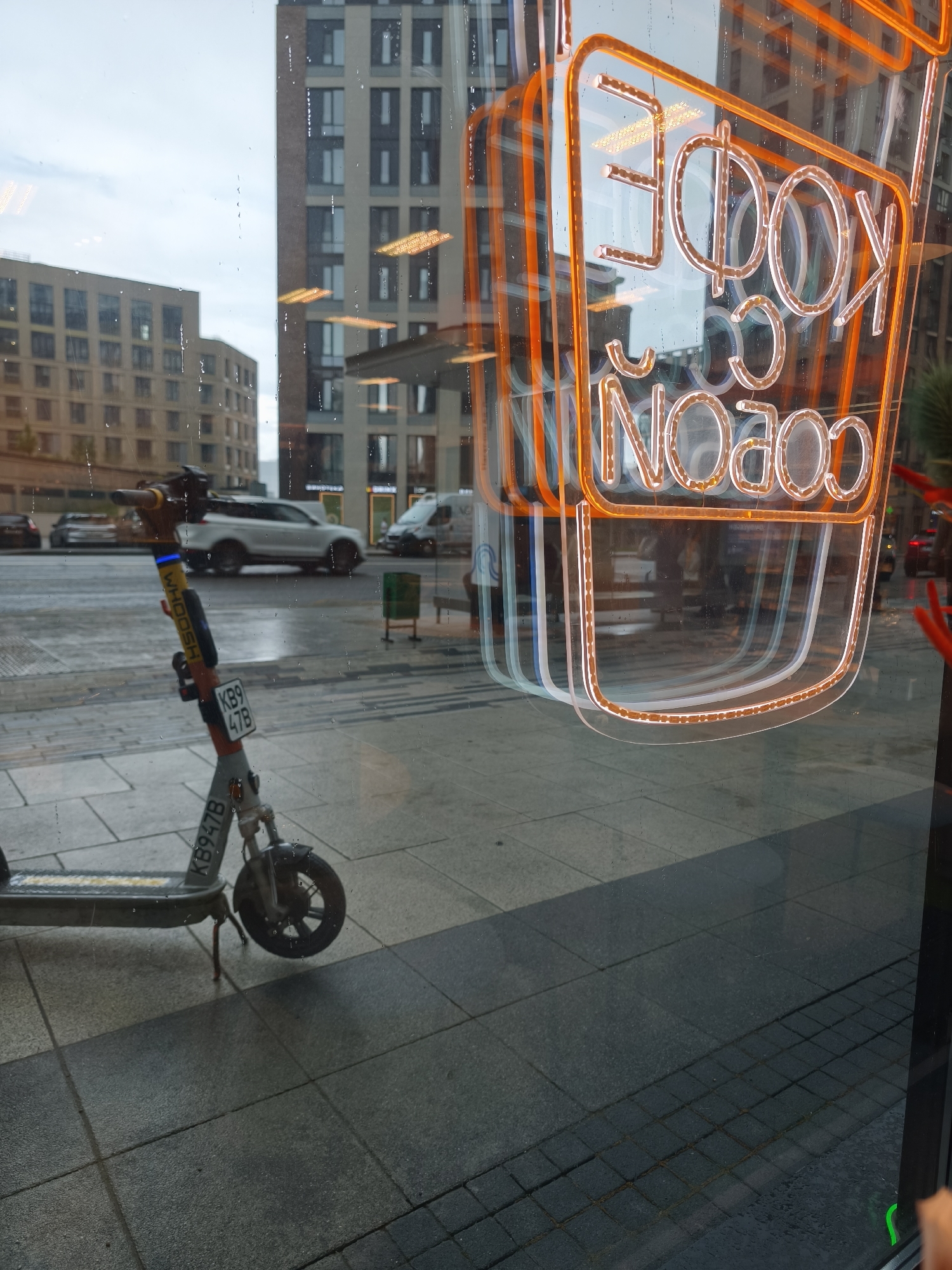 The image is a photograph taken through a window. Reflected in the window are urban elements: buildings of various heights with repetitive window patterns, street, and several cars parked or moving. A scooter is parked close to the window. The scooter is light grey and has a license plate reading "KB9 47B". A sticker on the front of the scooter reads "WHOOSH". The street appears wet. The window has condensation spots. Overlaying the reflection is an orange neon sign in Cyrillic, reading "КОФЕ С СОБОЙ" (Coffee To Go). The image appears to be taken in Moscow, Russia, given the Cyrillic text.