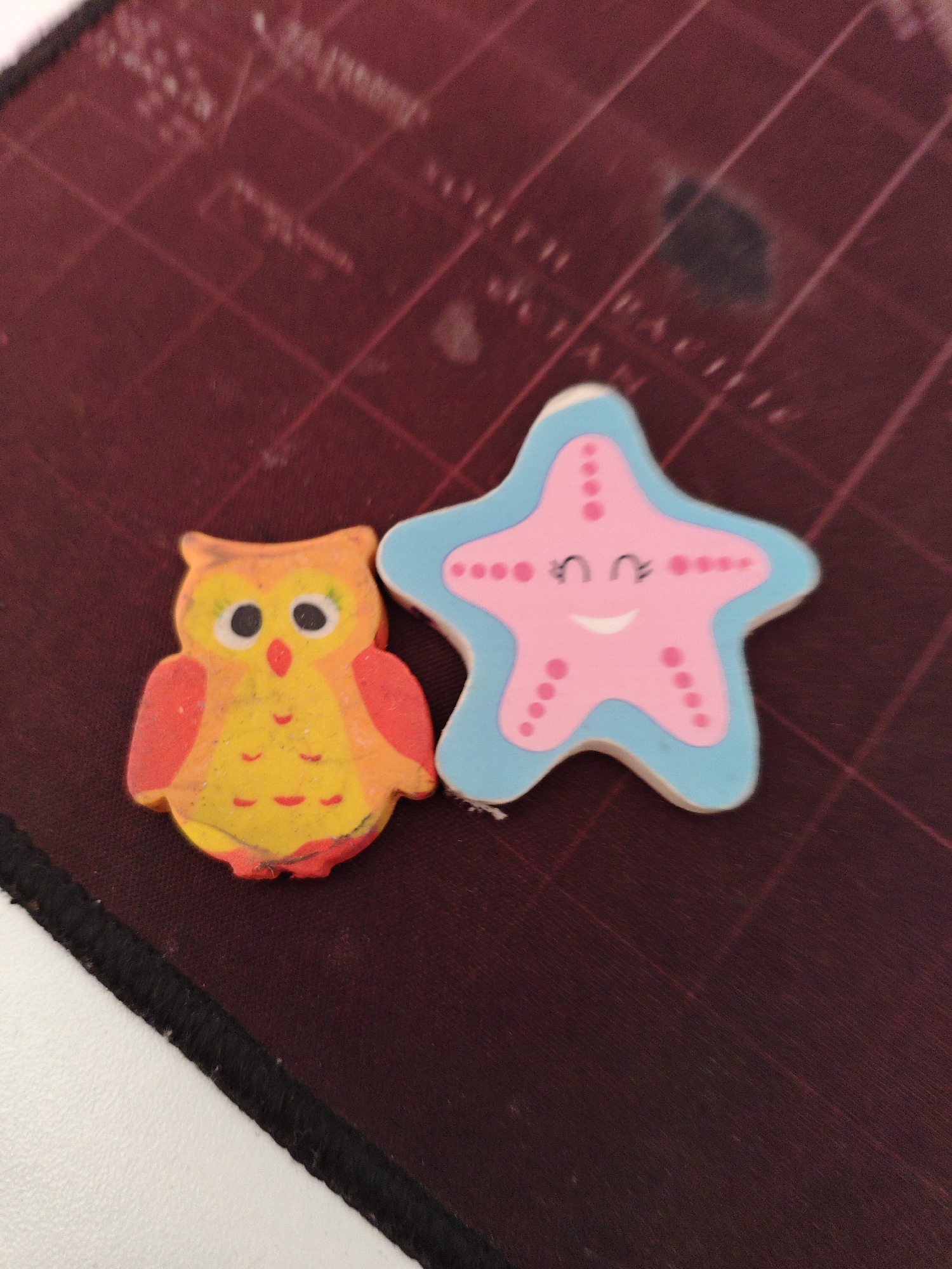 Two novelty erasers, shaped as an owl and a starfish, rest on a dark red, gridded surface, likely a cutting mat. The owl eraser is primarily yellow with red accents on its wings, eyes, and beak. The starfish eraser is pink with a light blue outline and features a smiling face with two black eyes and a curved mouth, along with several small pink dots representing its texture. The background surface has faint grid lines and appears to have some smudges or scuffs. The lighting is moderate, casting soft shadows. The scene is a close-up shot, emphasizing the two erasers.