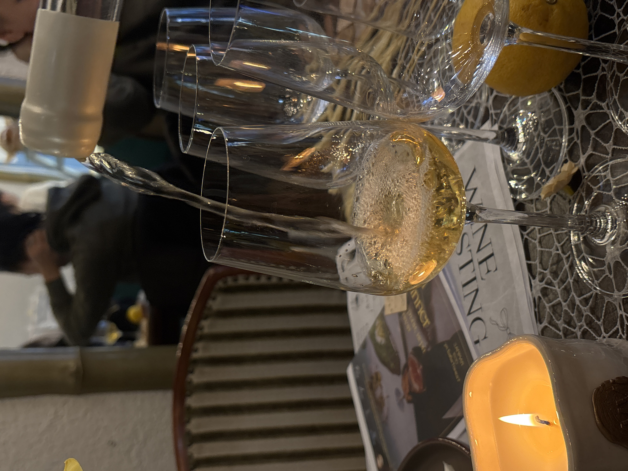 The image captures a still life scene on a table, likely indoors, featuring several champagne flutes, one of which is filled with a sparkling golden liquid. The flutes are arranged haphazardly, some empty and some overlapping. Next to the flutes is a lemon and a lit candle in a white, textured holder with a gold accent. A newspaper with the word "Tasting" is also visible.
The image also includes a reflection of a person in the background, and wooden chair.
There is no information to suggest that this image was taken in Varna, Bulgaria.