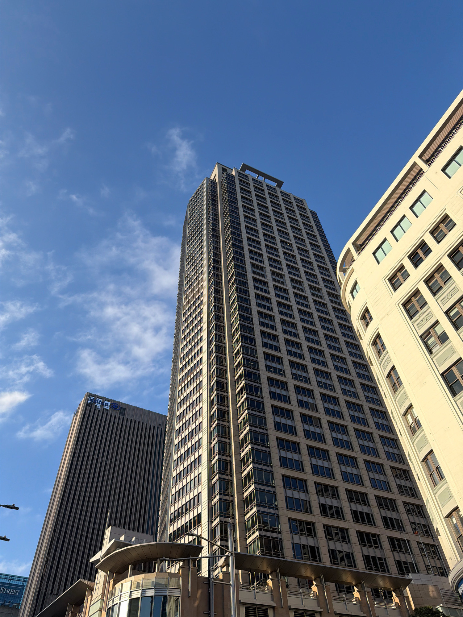The image shows a low-angle shot of tall modern buildings against a bright blue sky with scattered white clouds. The buildings are made of glass and concrete, with a repetitive pattern of windows. The central building is the tallest and appears to be a skyscraper with a dark facade. To its right, a lighter-colored building has a curved facade with visible balconies. To the left of the central building, another, slightly shorter building with a dark ribbed facade is visible. The scene is set in a city, likely a business district given the architecture. One of the buildings on the left has a sign with blue Korean characters that translates to "KB Kookmin Bank". The scene is captured on a sunny day, suggesting good weather. The perspective is from street level, looking upwards towards the tops of the buildings.