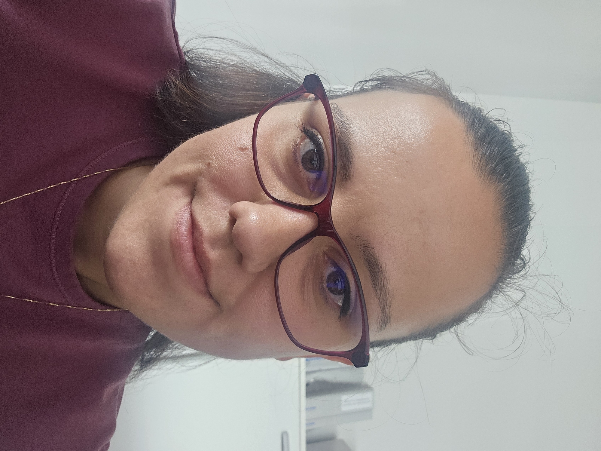 A close-up, chest-up portrait of a woman wearing dark red-rimmed glasses and a burgundy sweatshirt. The woman has dark hair pulled back and a small mole on her left cheek. She is looking directly at the camera with a slight smile. The background is a plain, light-colored wall with some blurred office-like shelves and cabinets visible on the right. The image appears to have been taken indoors, likely in an office environment in Bozhurishte, Bulgaria.