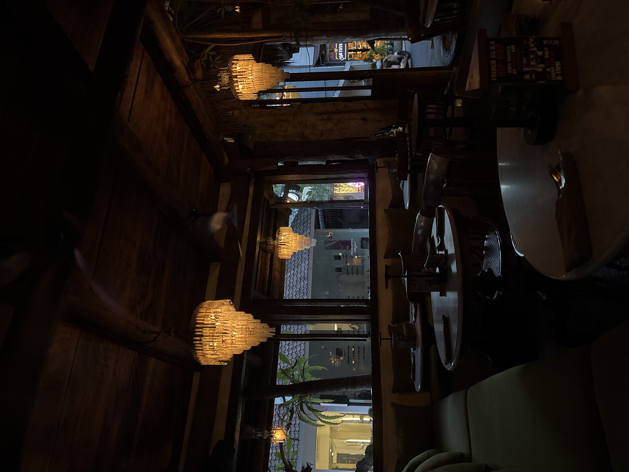 The image depicts an indoor space in Mengwi, Indonesia. The view looks through what seems to be an open-air restaurant towards an exterior street. Inside, there are tables and seating, including what appears to be a green couch. Hanging from the ceiling are ornate, multi-tiered light fixtures. The interior has a rustic feel, with wooden beams and walls. Looking out from the restaurant, the streetscape includes other shops or buildings with signage. The time of day seems to be evening or night.