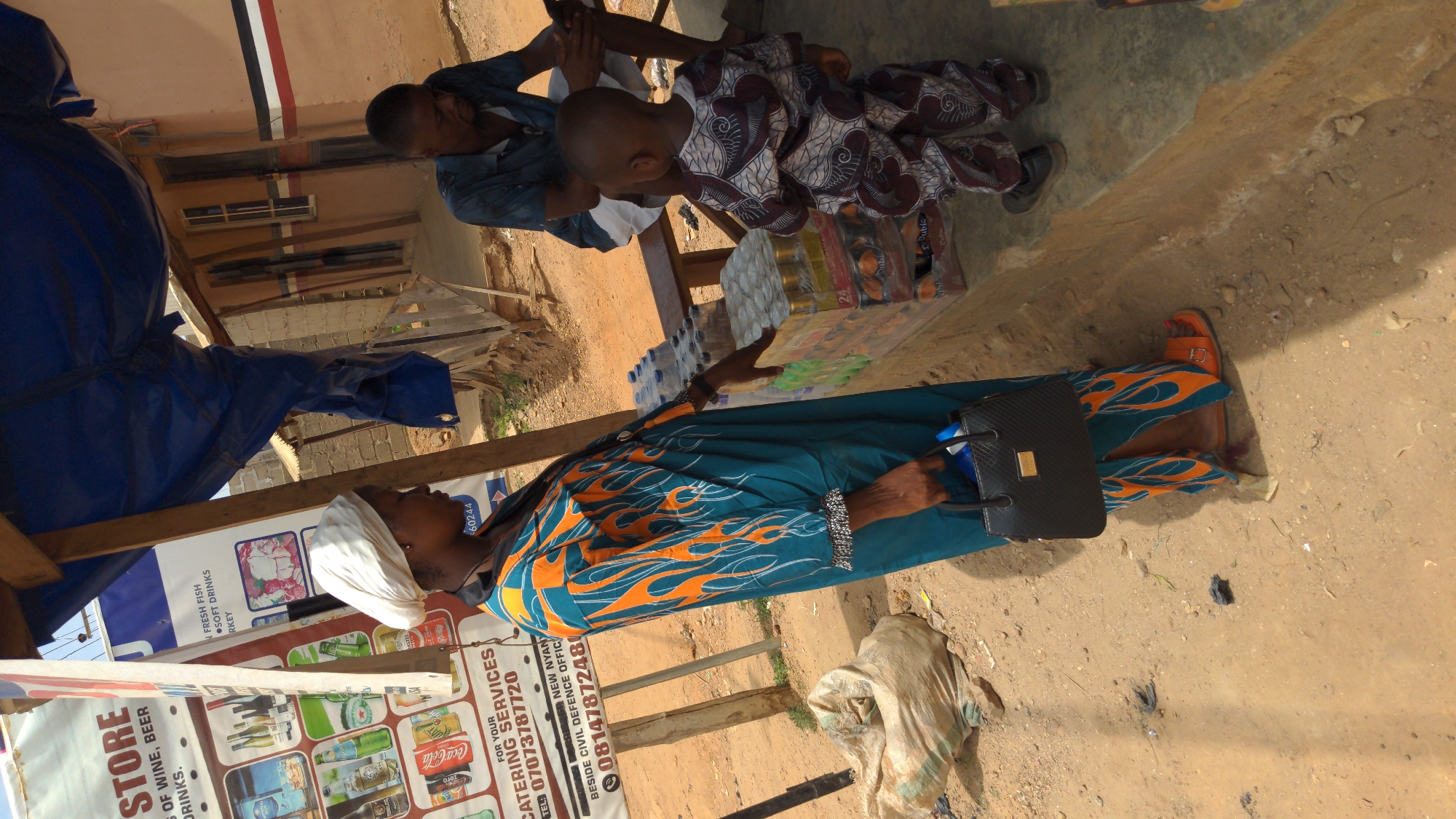 The image shows a street scene in Ado, Nigeria. An adult woman, dressed in a turquoise and orange patterned gown and wearing a white head wrap, is seated at a table, holding a package of what appear to be eggs and a small black handbag. To her left is a sign advertising "Catering Services" with a phone number and stating "Store: Of wine, beer drinks," alongside images of various beverages and food items. To the right of the woman, a man and a child are seated on a bench near bottles of water and other packaged goods. The ground is sandy.