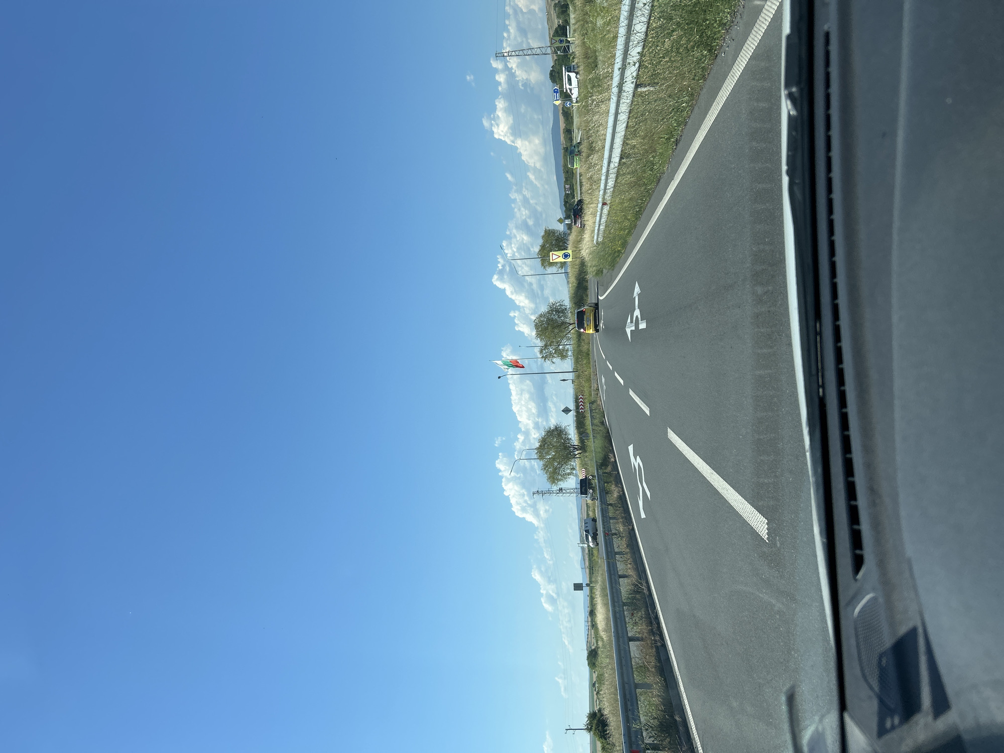 The image captures a view from inside a vehicle driving on a highway near Aheloy, Bulgaria. The sky is a clear, bright blue with scattered white clouds. A paved road stretches forward, marked with white lane lines and directional arrows indicating a right turn and straight ahead. Several vehicles are visible on the road, including a yellow car, a dark SUV, and a white van. A metal guardrail lines the right side of the road, with dry grass and vegetation growing alongside it. In the distance, there are rolling hills and trees. A Bulgarian flag is prominently displayed on a flagpole to the left of the road. The dashboard of the vehicle is visible in the foreground.