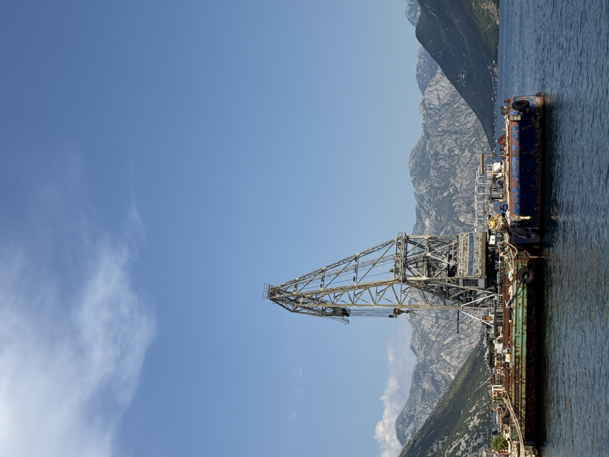 The image shows a clear blue sky with a few wispy clouds above a body of water. In the foreground, a large industrial crane stands on a barge, its metallic structure weathered and rusted. The barge itself appears old and worn, with various pieces of equipment and debris scattered across its deck. Behind the barge, rugged, mountainous terrain rises up from the water, covered in sparse vegetation. The overall scene suggests a working port or industrial area situated in a naturally dramatic landscape. The location is identified as Lipci, Montenegro. There are no people visible in the image.
